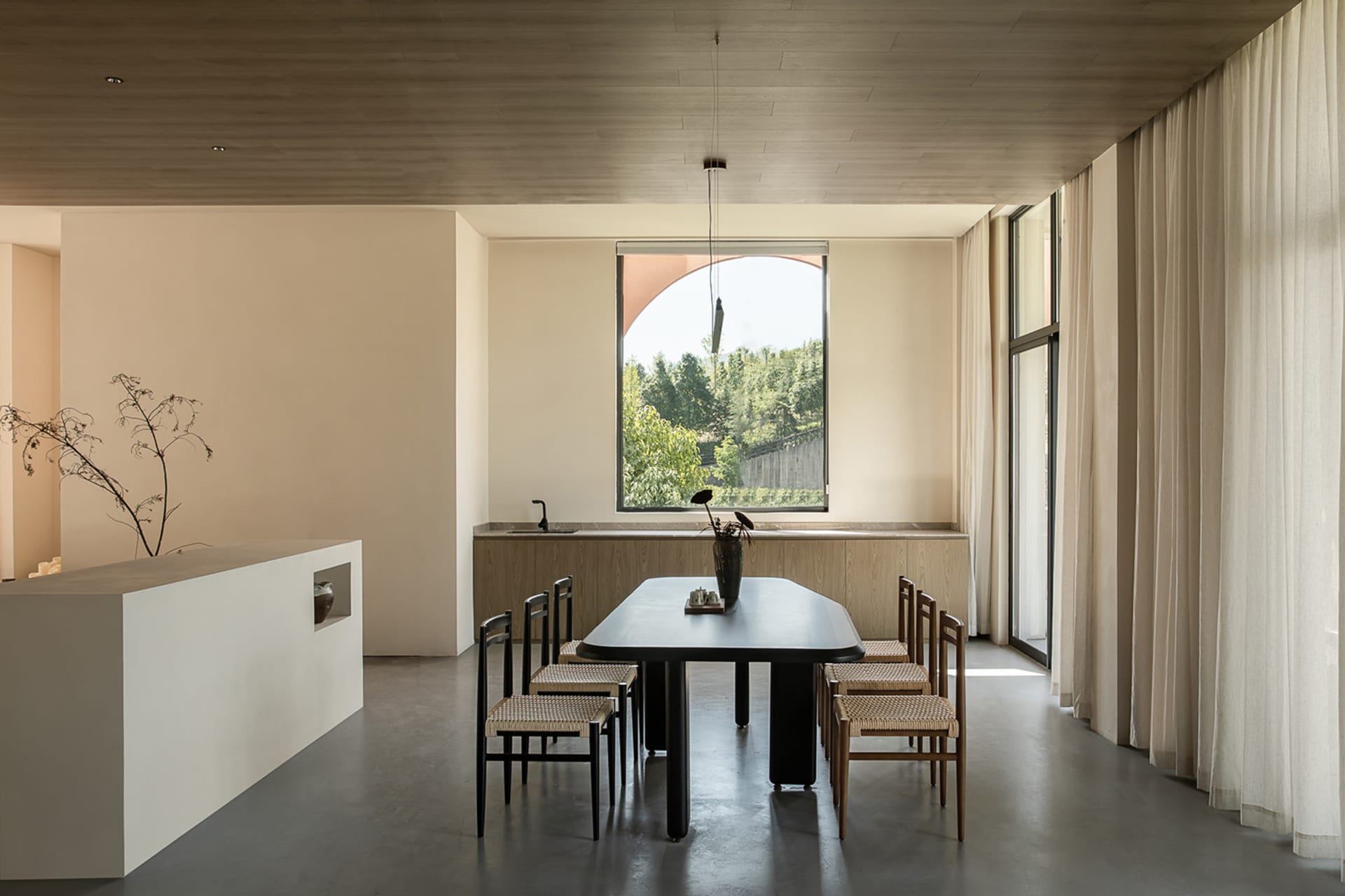 Modern dining room with wooden furniture, a leaning flower pot, and a view of greenery outside.