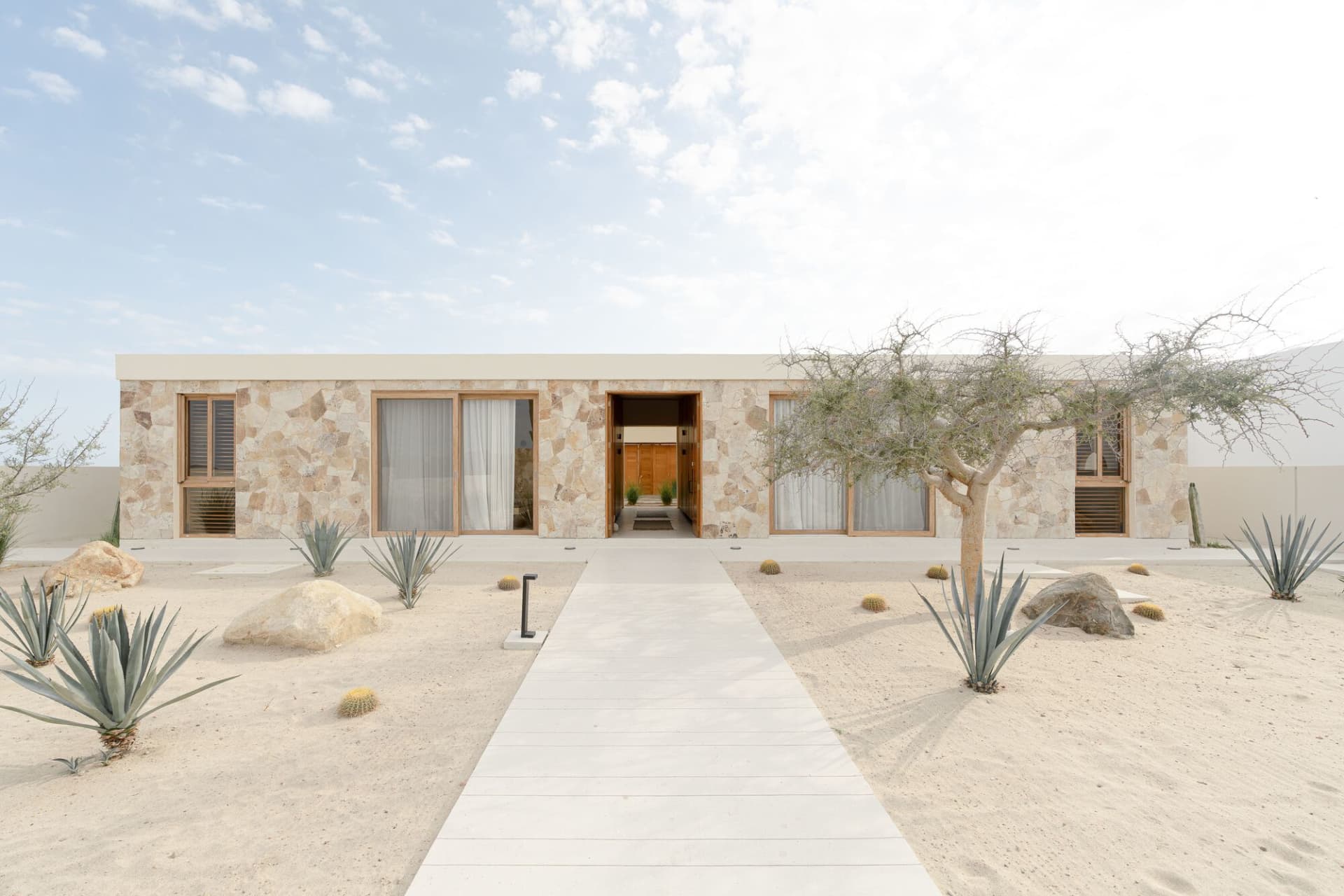 A modern minimalist residential house with a flat roof and large windows, constructed from sandstone and featuring cactus and