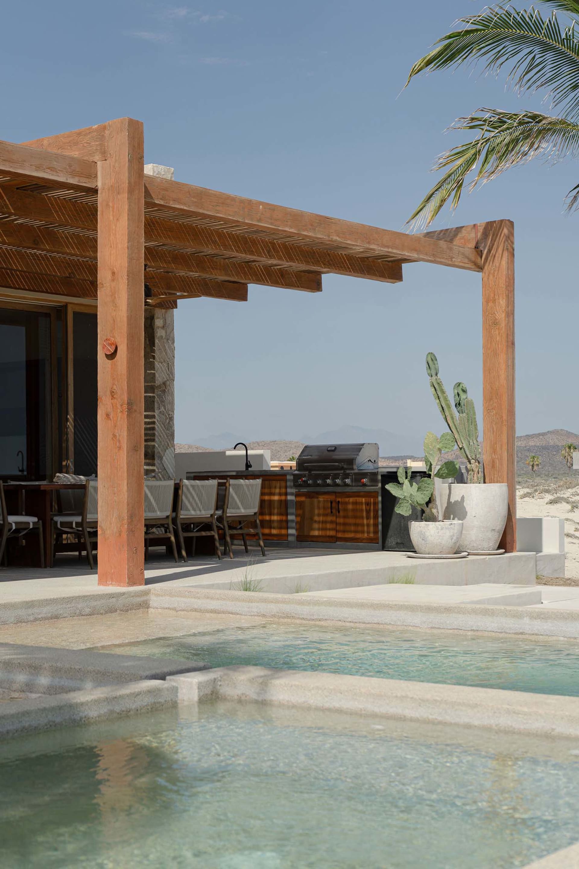 Modern residential pool area featuring a pergola-covered outdoor kitchen, grill, dining table, and cactus plants.