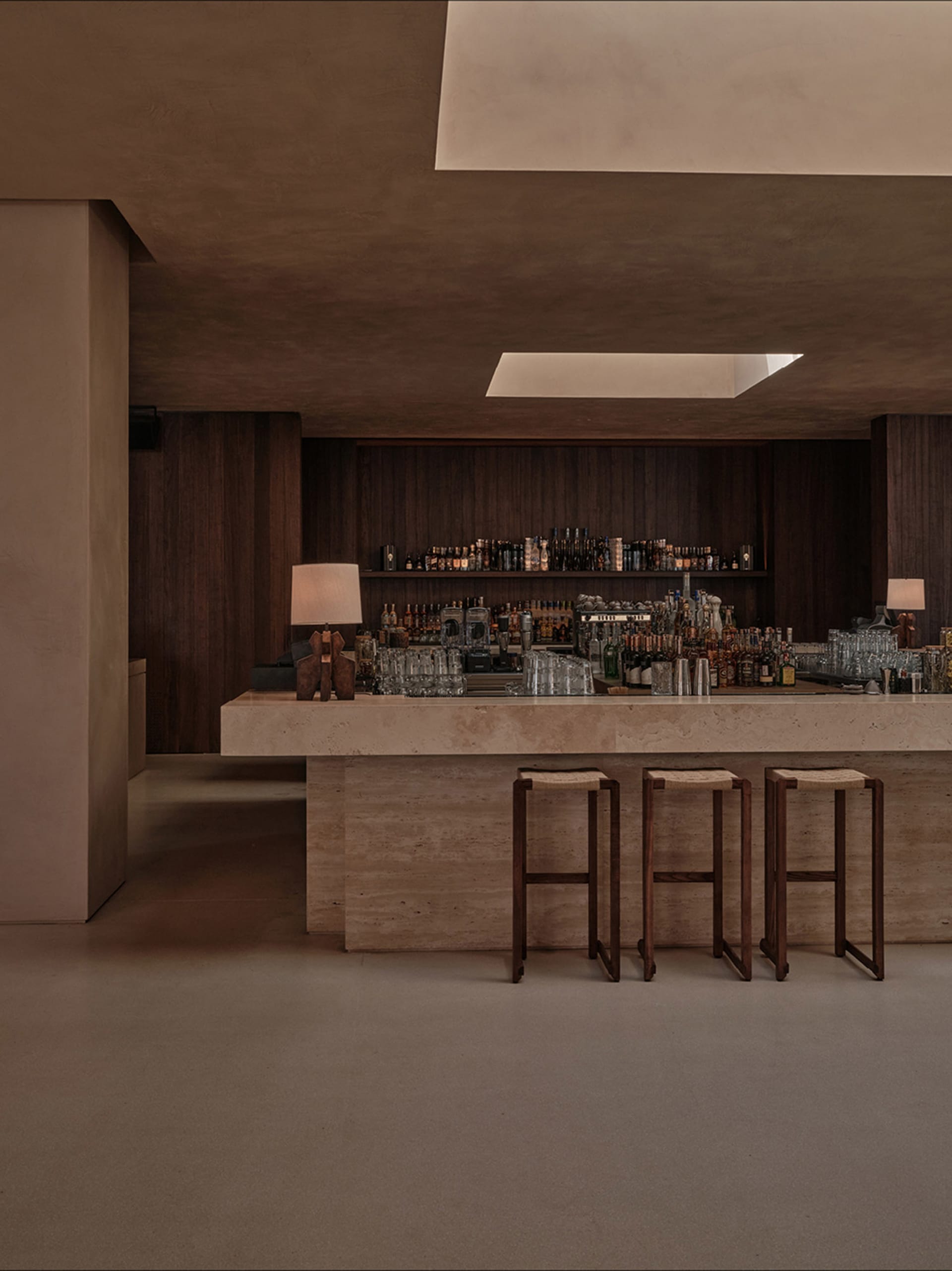 A minimalistic bar area with a stone countertop, wooden stools, and glassware on shelves, illuminated by warm lighting.
