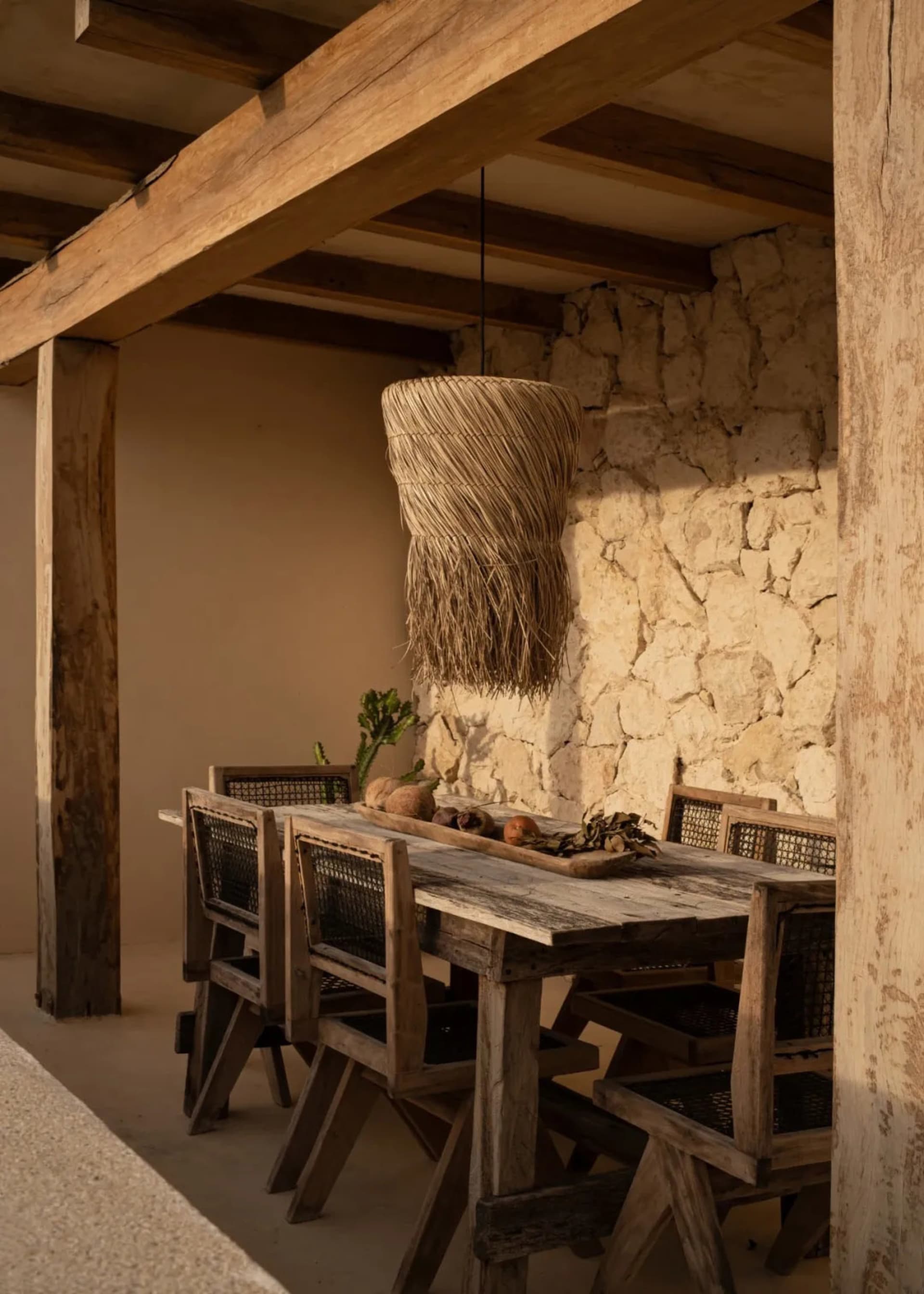 A rustic farmhouse dining room with wooden tables, chairs, and a woven pendant light hanging from a wooden beamed ceiling.