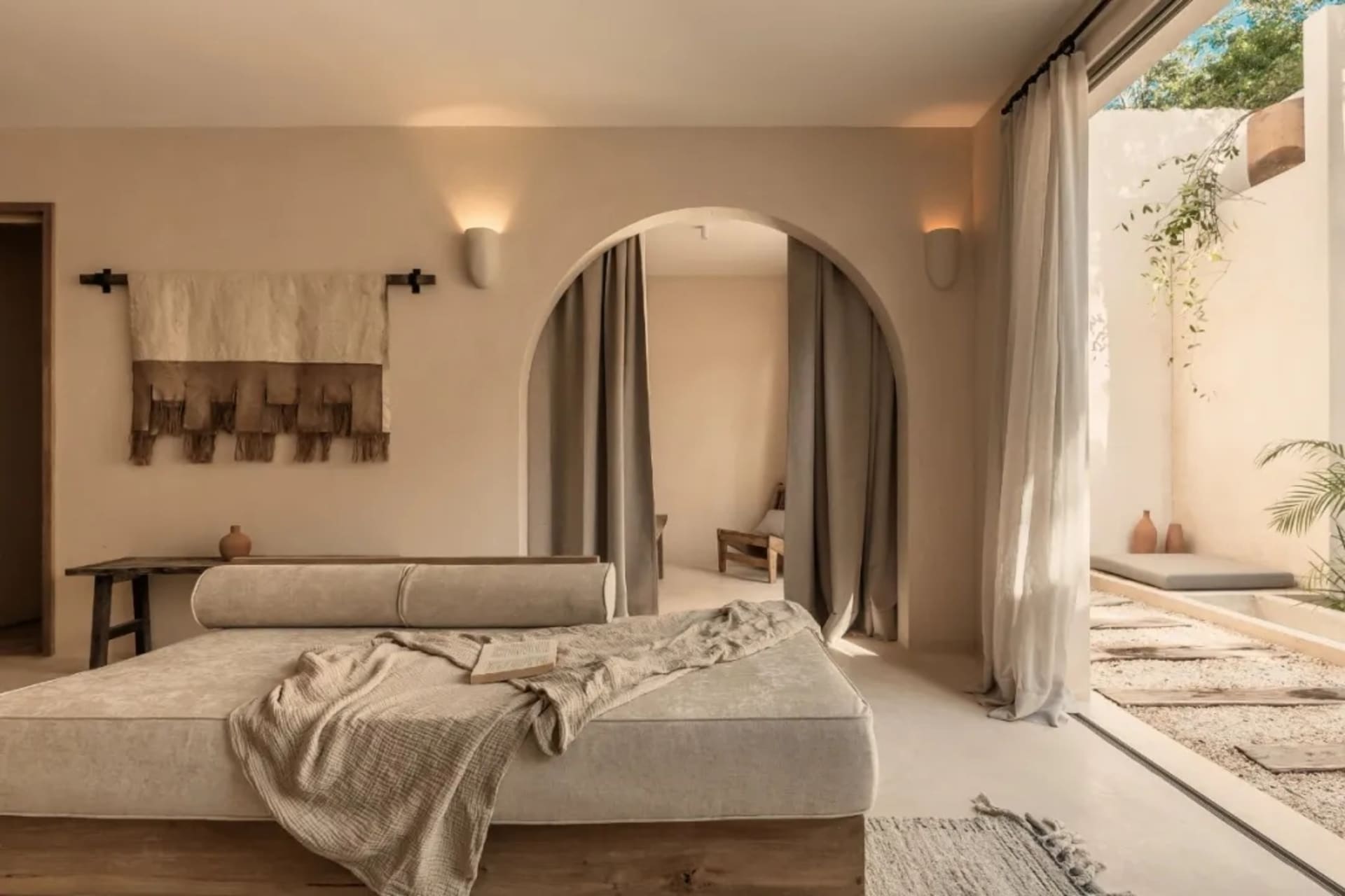 A minimalist bedroom featuring a bed with fabric bedding and curtains, surrounded by neutral tones and natural elements.