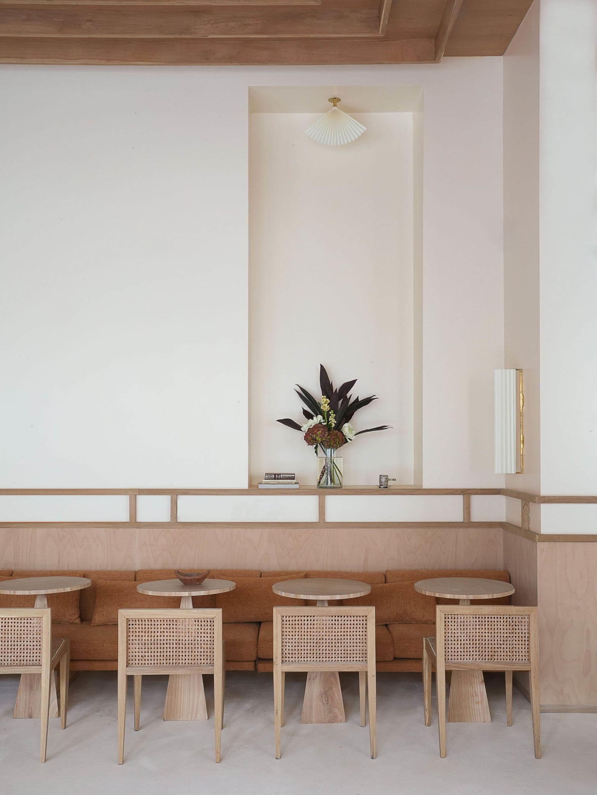 A modern cafe interior featuring a long wooden sofa, round tables, and cane-back chairs.
