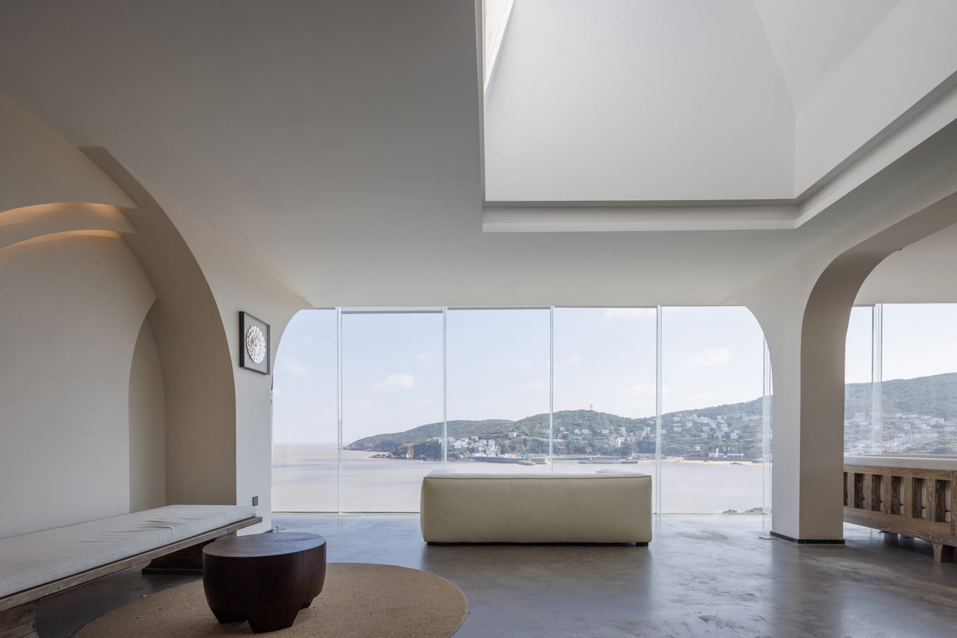 Modern living room with large glass windows overlooking the ocean, featuring sleek furniture and minimalist decor.