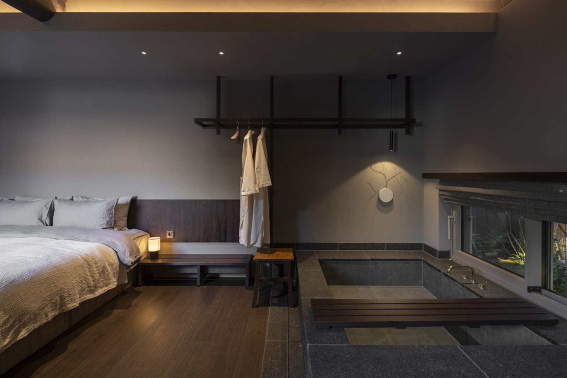 A minimalist bedroom featuring wood flooring, a stone bathtub, and a bed with textiles.