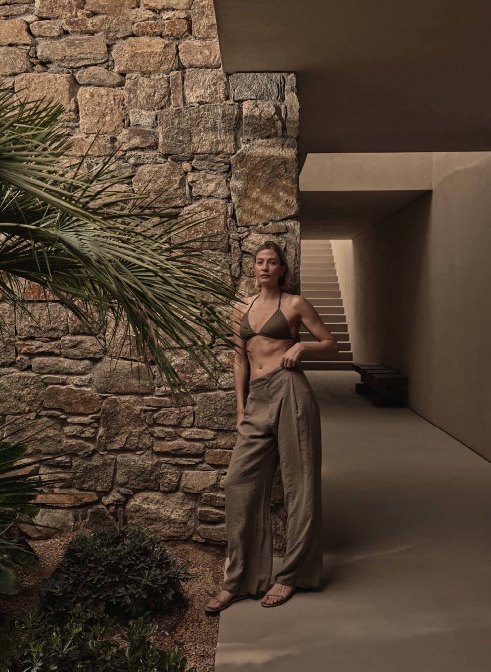 A woman wearing a minimalist outfit, including a bikini top and wide-leg pants, stands near a stone wall.