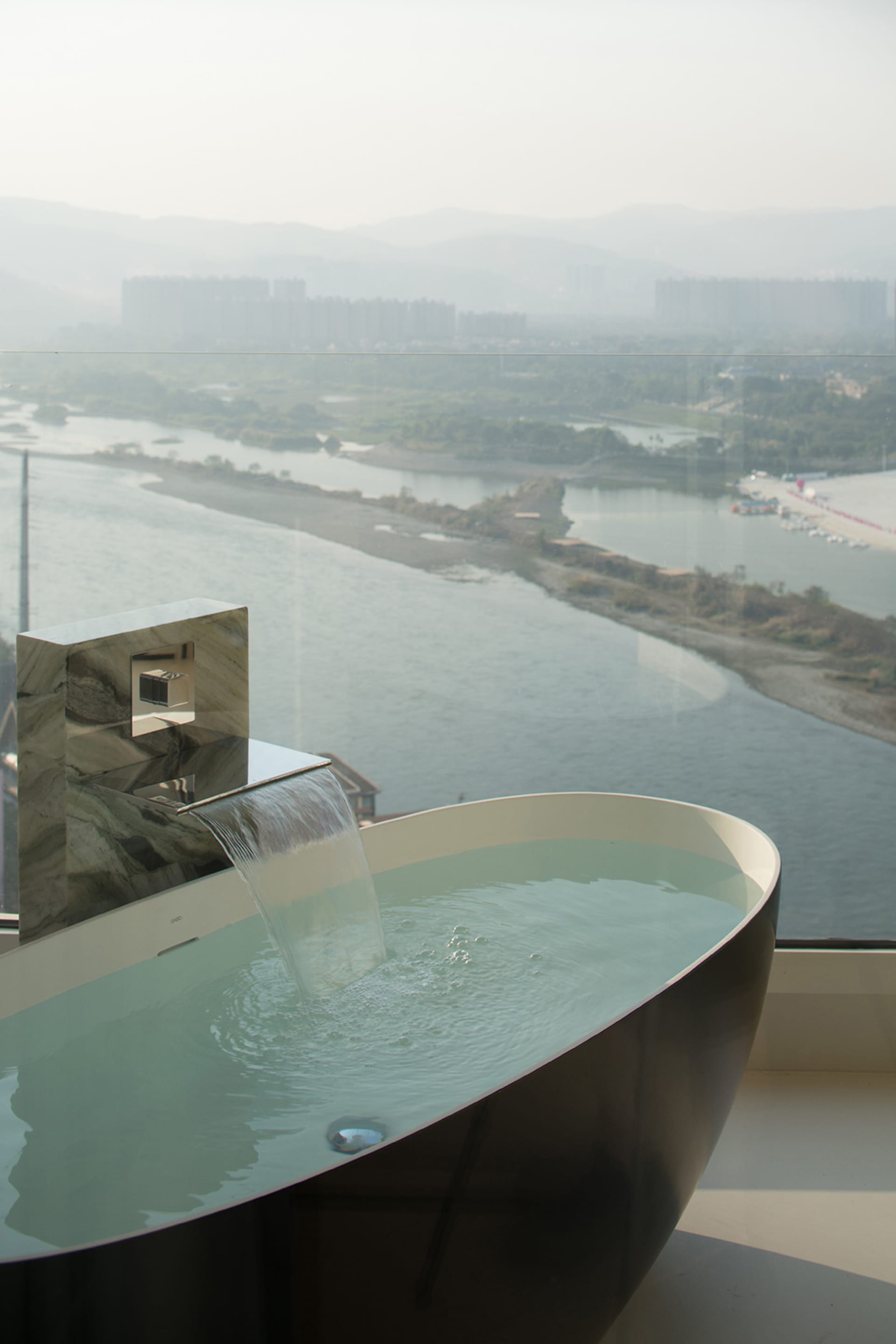 Modern bathroom with glass waterfall bathtub overlooking scenic river and cityscape.