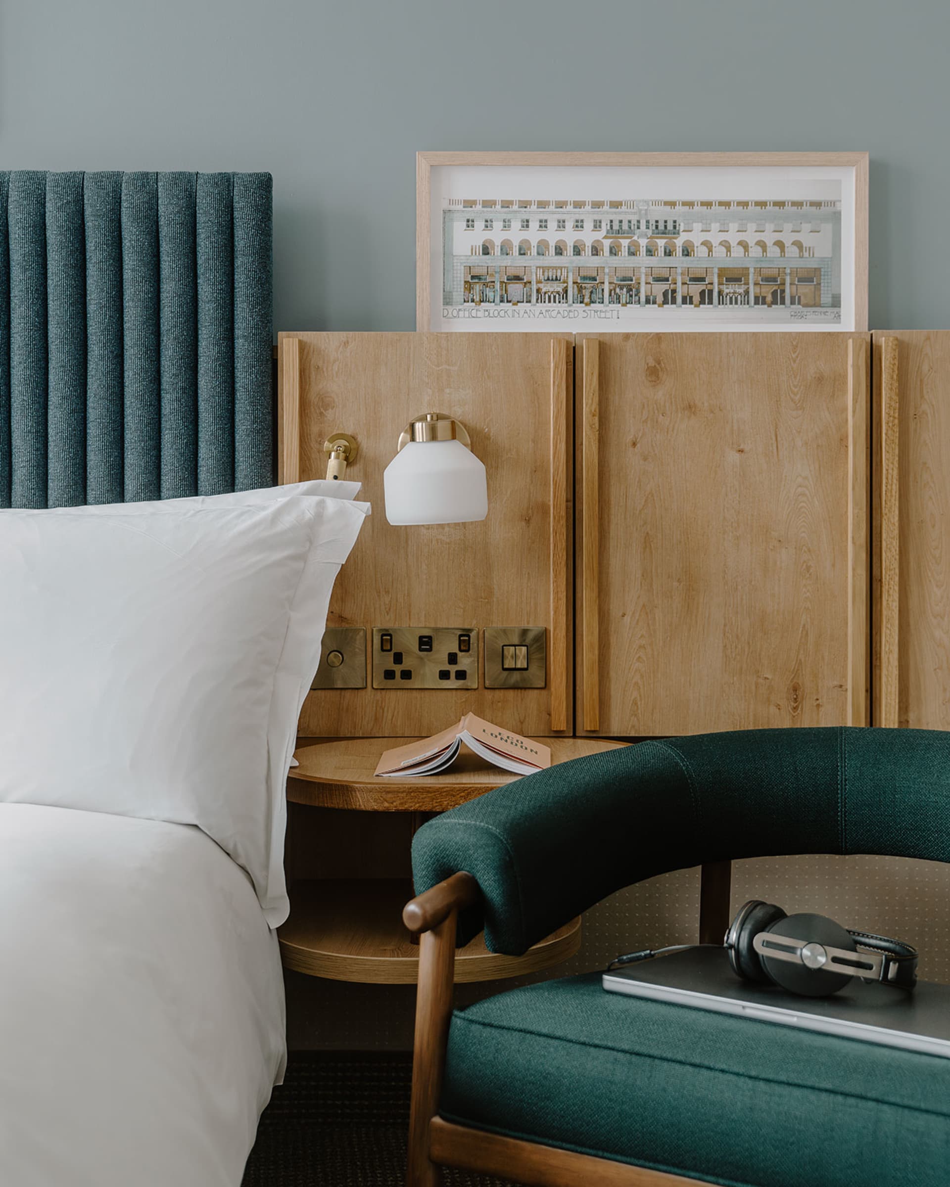 A minimalist hotel room featuring a bed with a wooden headboard and a wooden nightstand with a lamp, a chair with a wooden ar
