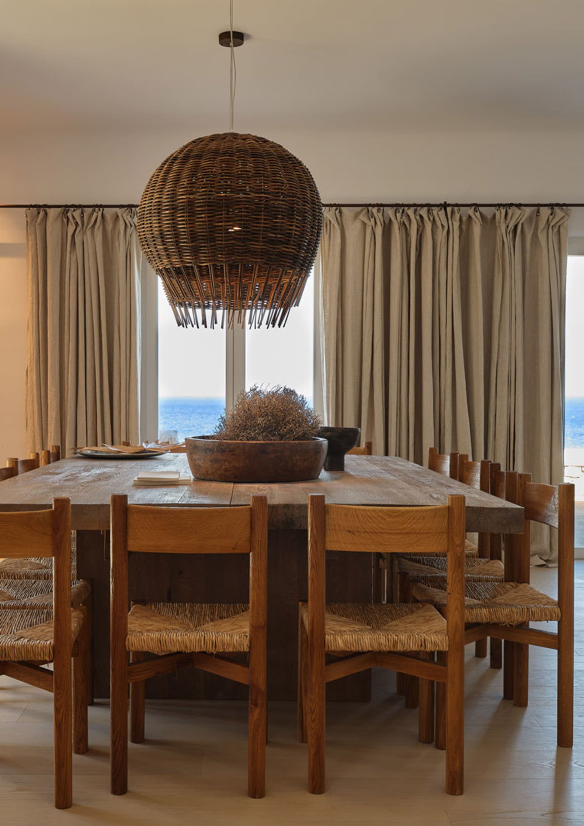 A minimalist dining room features a wooden table and chairs, with a wicker chandelier hanging overhead and curtains framing a
