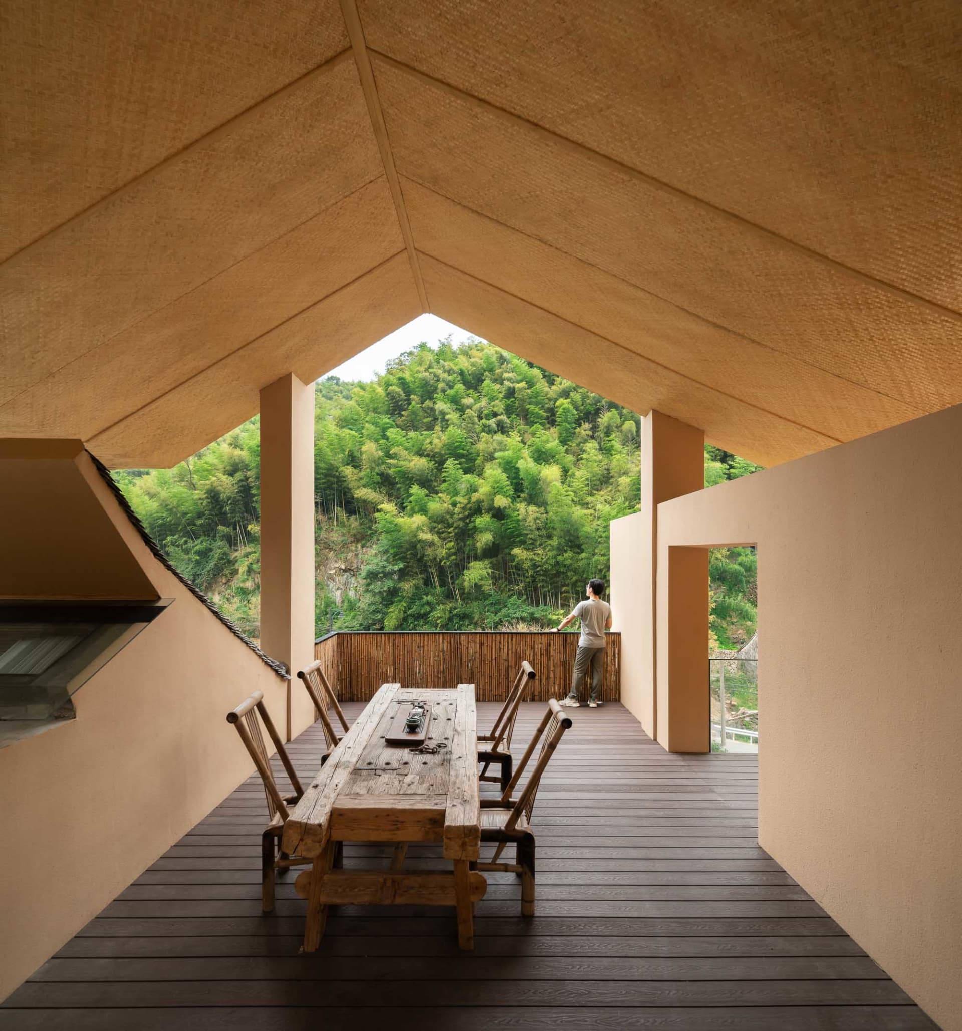 A contemporary residential exterior featuring wood and bamboo materials, with a person standing on a wooden deck overlooking 