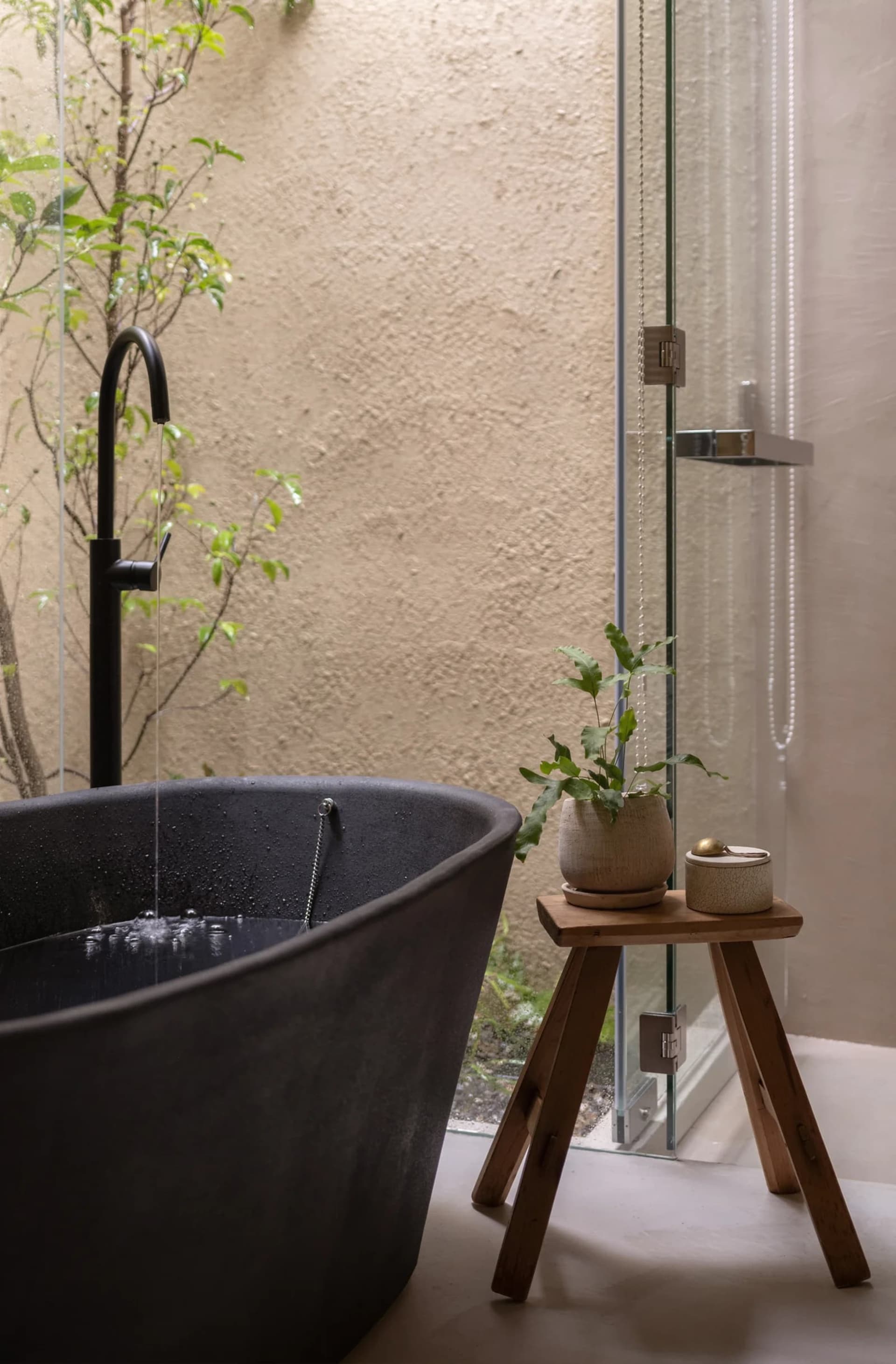 A modern, minimalist bathroom with a freestanding concrete bathtub, a small wooden stool with a plant and a container, and a 