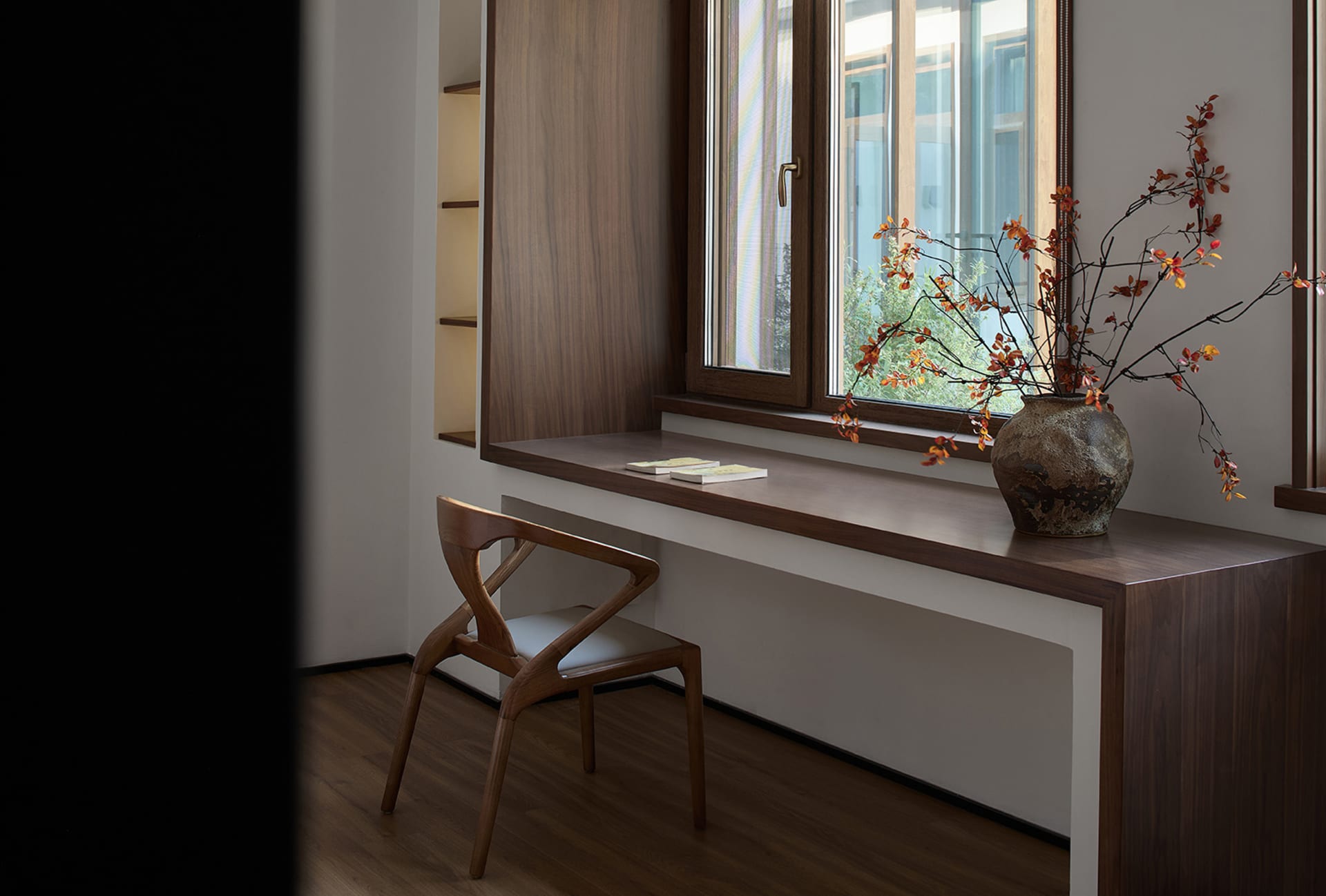 A modern library space featuring a wooden desk, chair, and bookshelves with a vase of autumn leaves on the windowsill.
