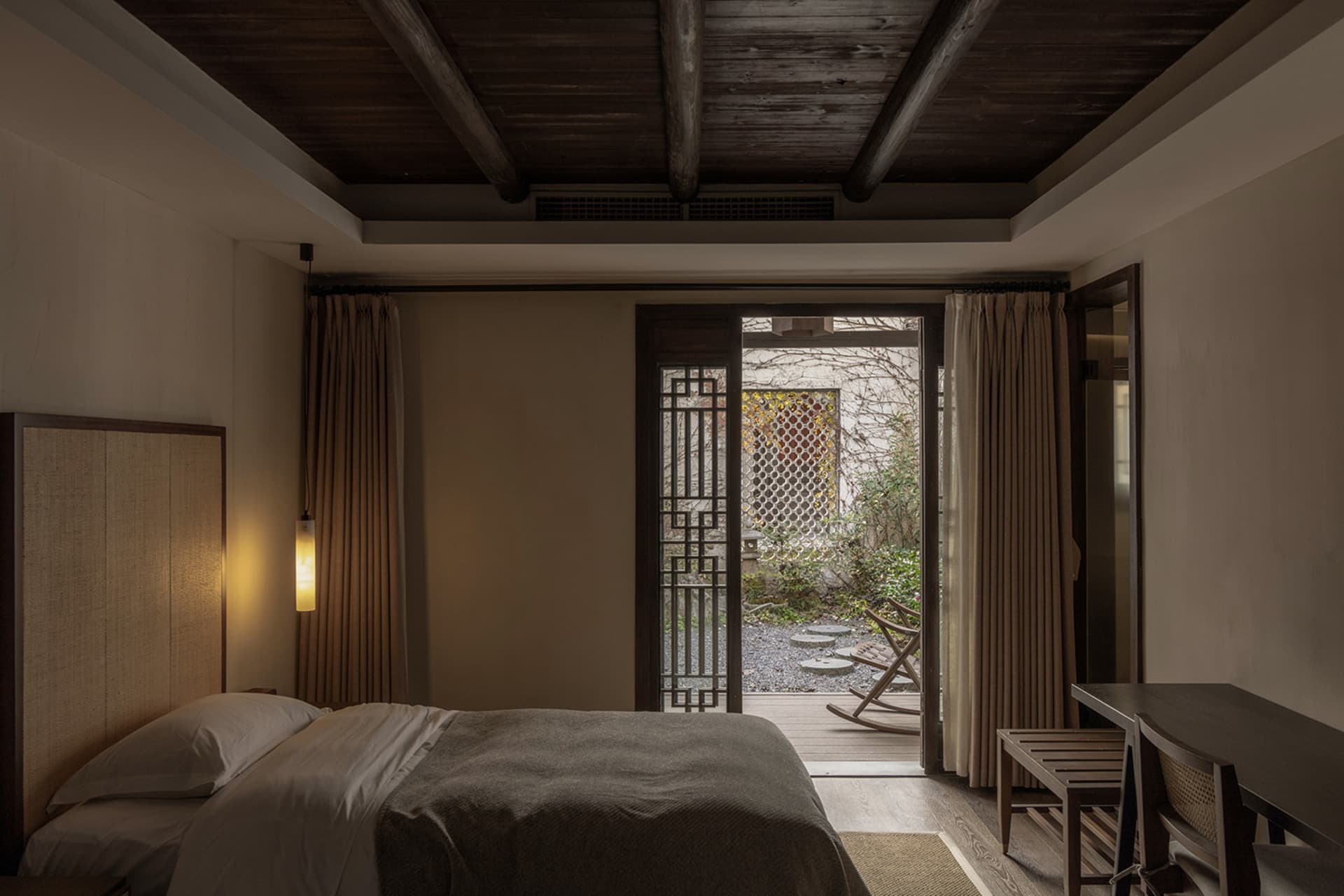 A serene minimalist bedroom with wooden and fabric elements, featuring a neatly made bed and a view of a serene outdoor area.