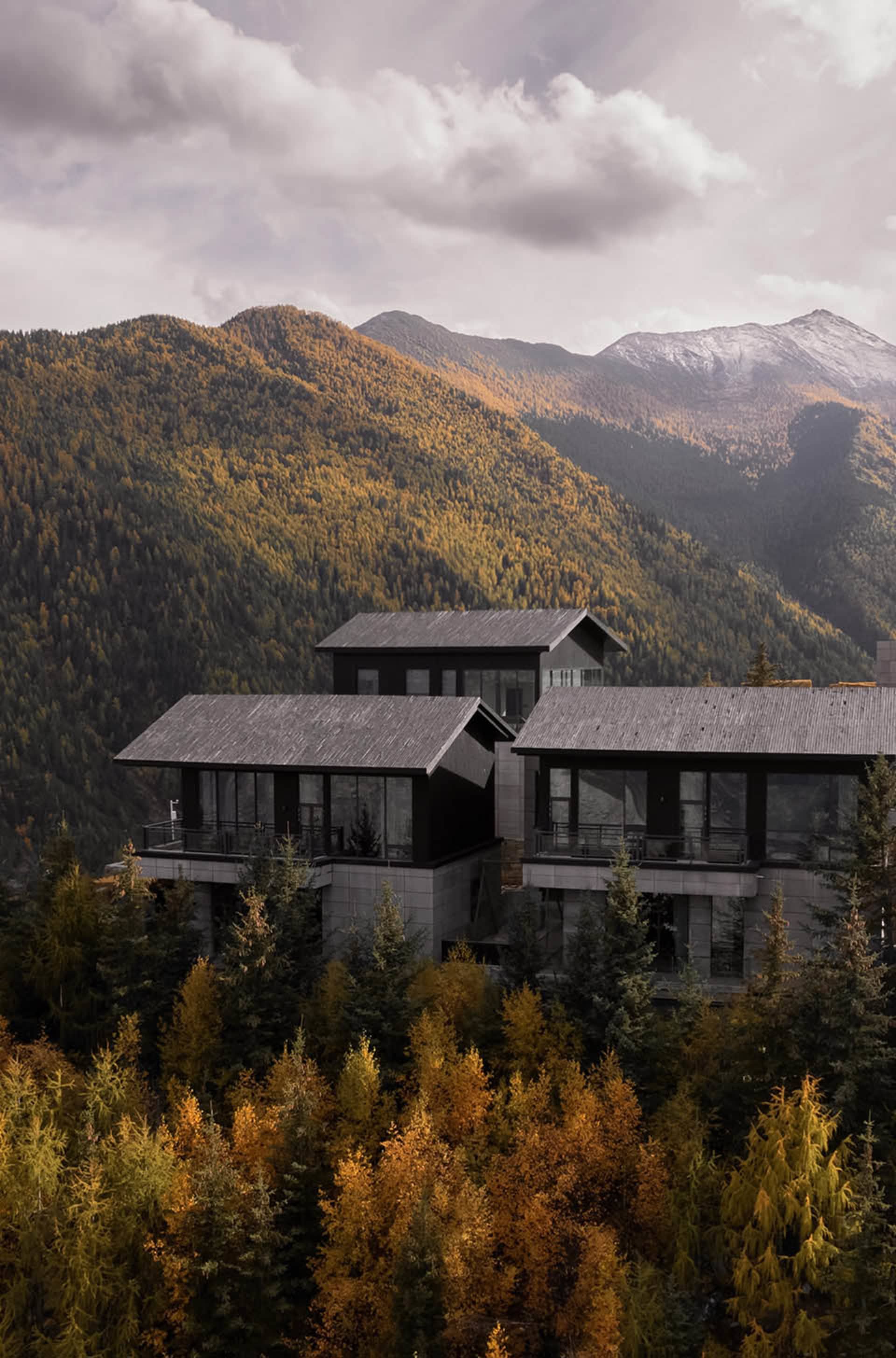 Modern, luxurious villa with black roofs nestled in a mountainous landscape surrounded by autumn-colored trees and foliage.