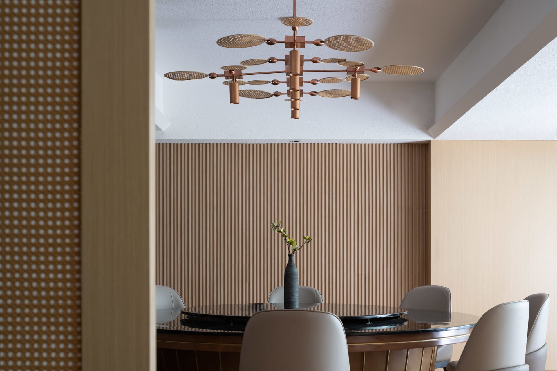 Modern dining room with a round table, leather chairs, and a vase of flowers, featuring a wood-textured wall and copper chand