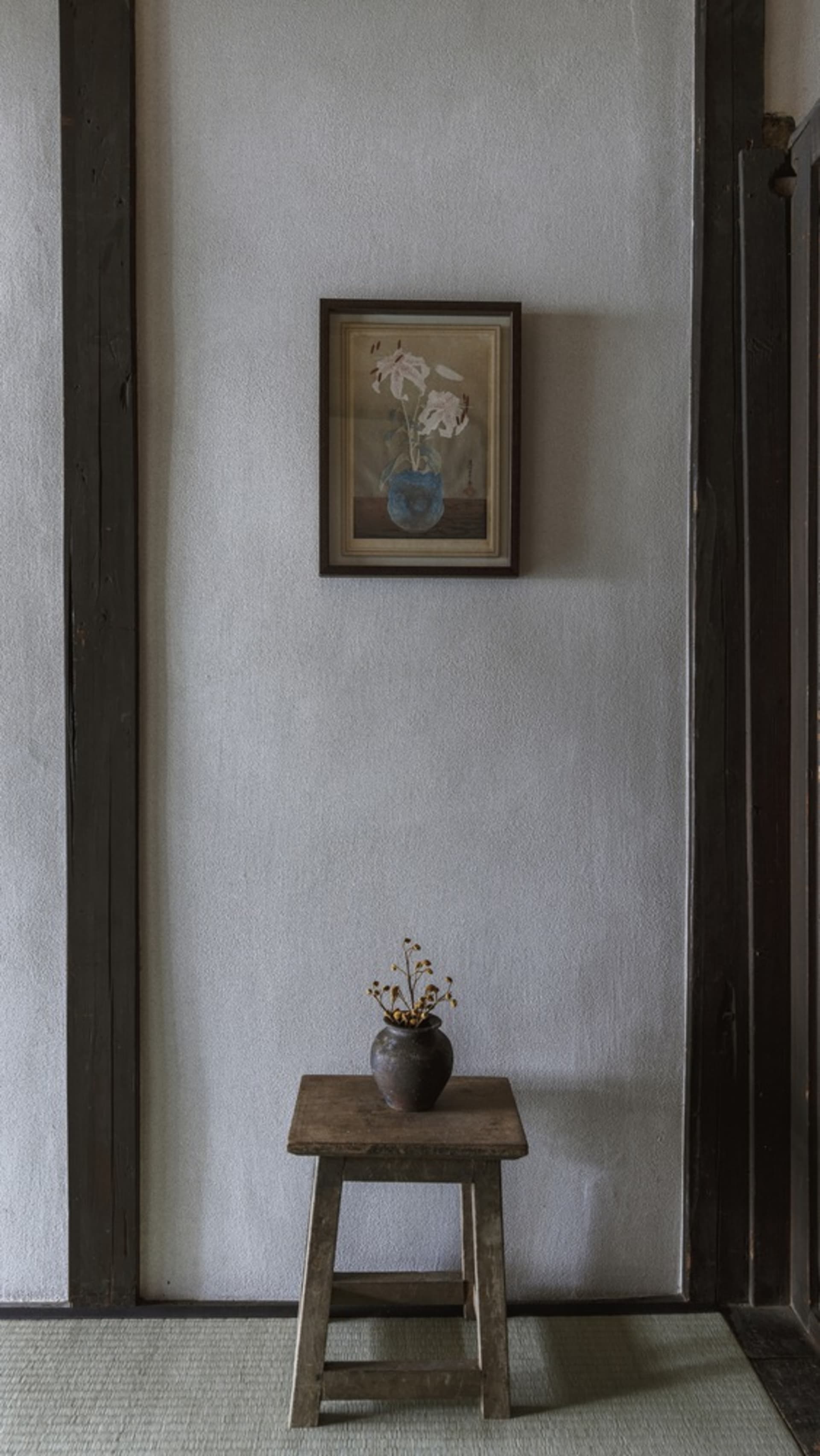 A minimalist studio featuring a small wooden stool with a vase of wilted flowers and a framed painting of flowers on the wall