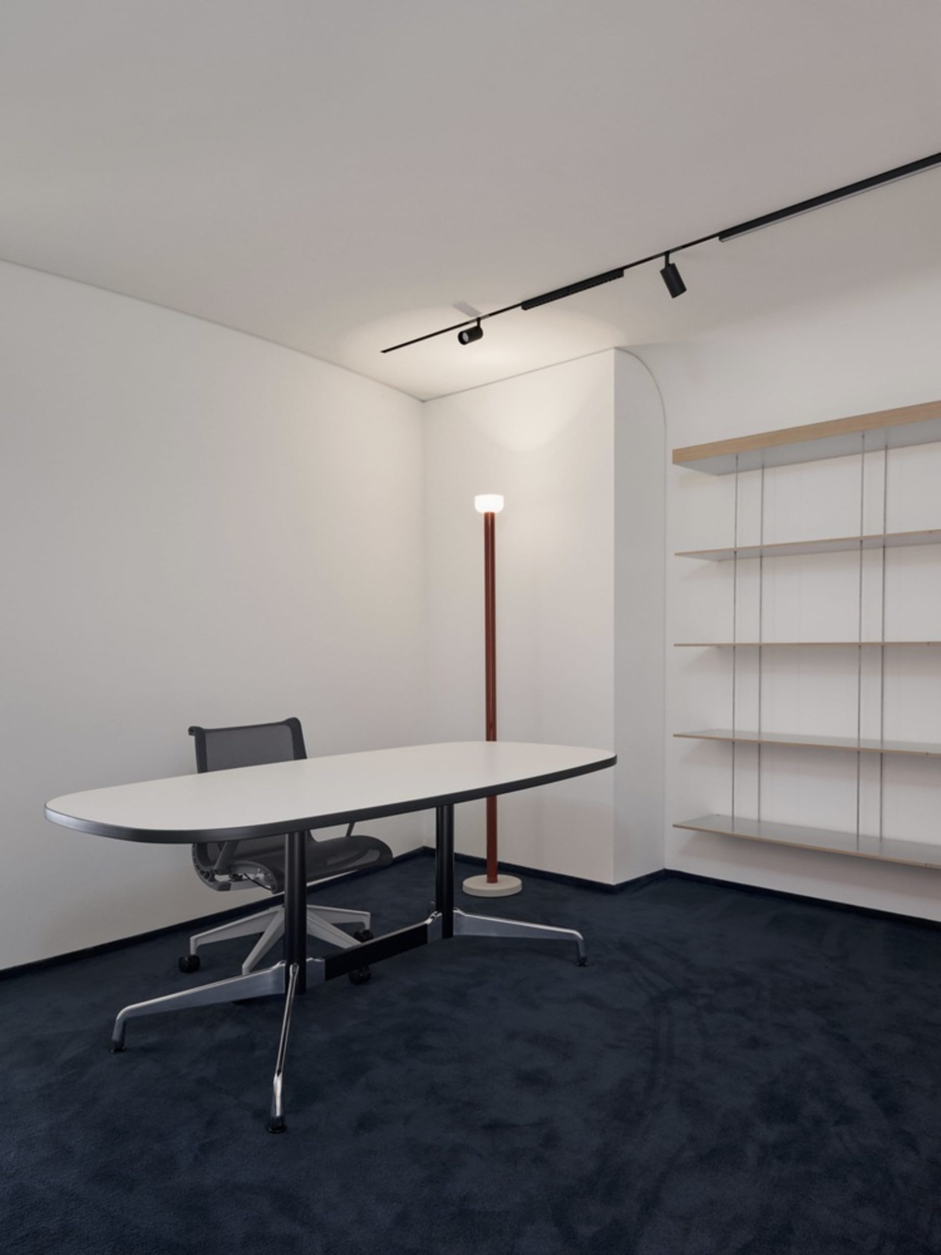A minimalist office featuring a white-painted wall, dark carpet floors, recessed lighting, and minimalist shelving.