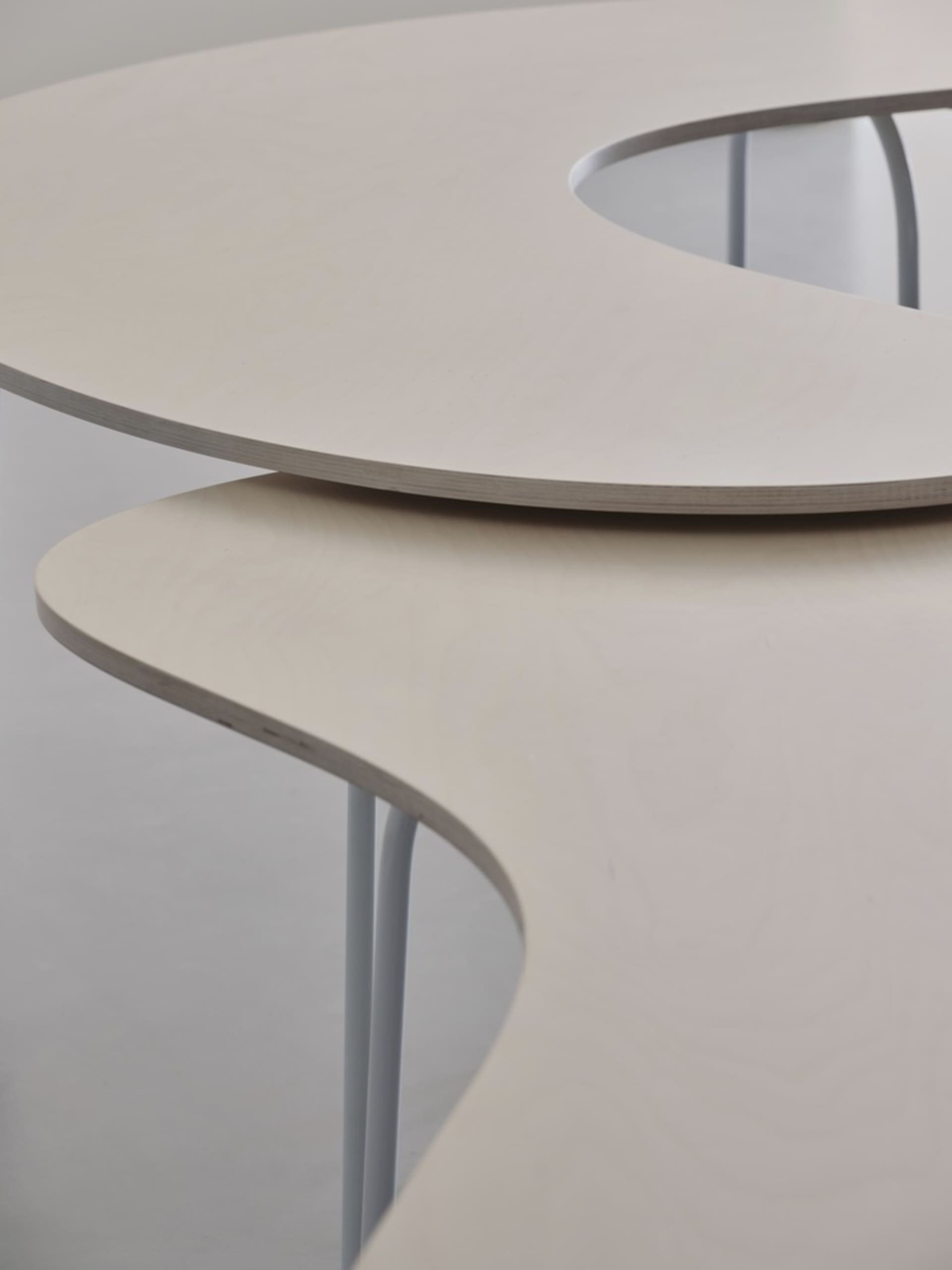 A modern furniture table with a curved, wooden tabletop and white metal legs.