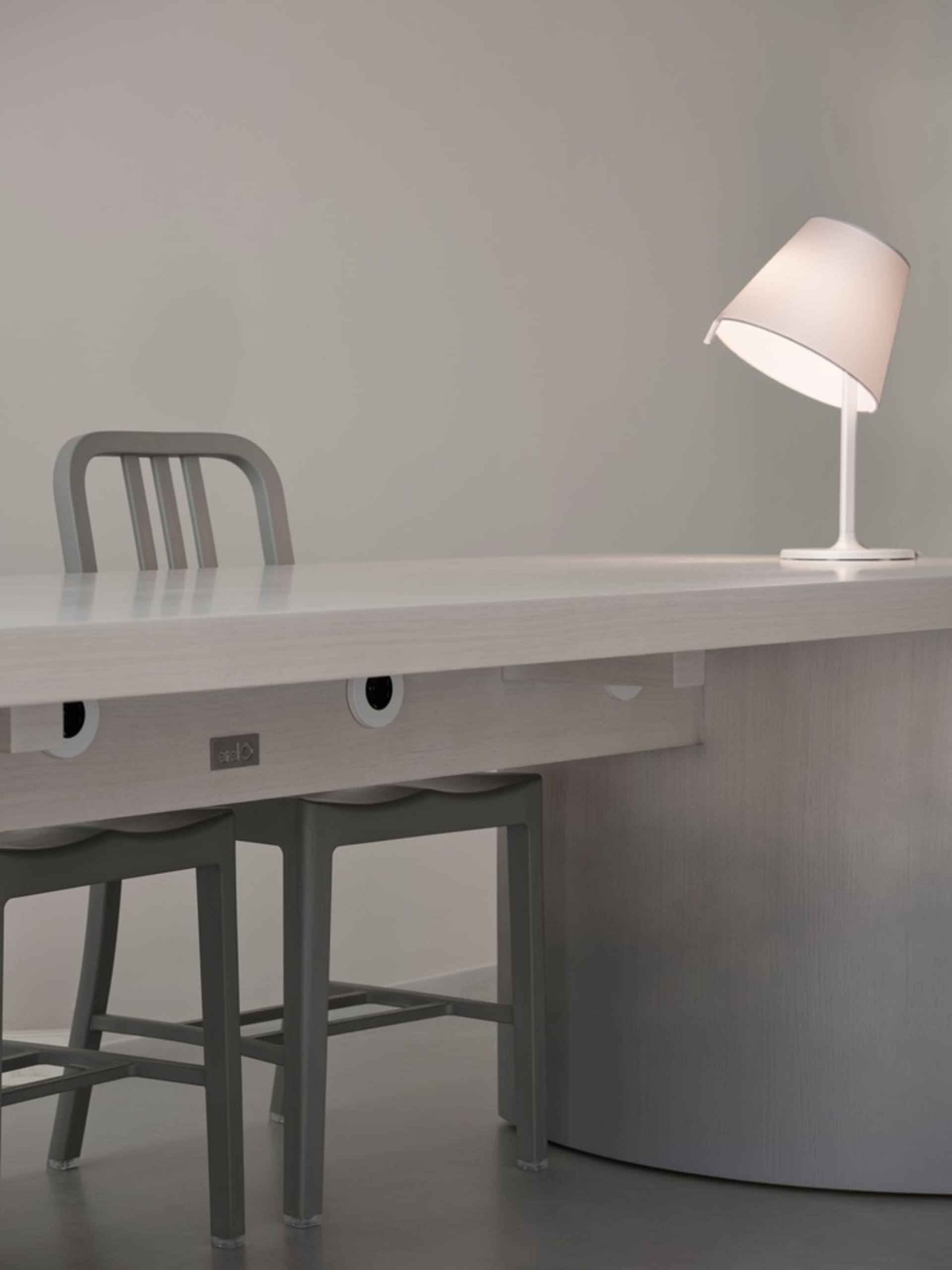 A minimalist office desk setup featuring a white desk, two stools, and a modern table lamp.