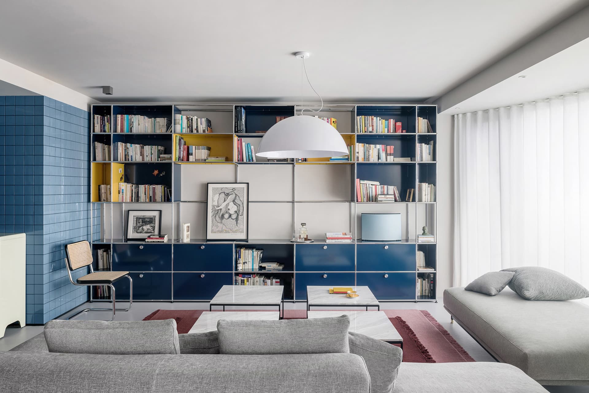 A modern living room with minimalist furniture, a metal and glass bookshelf, and fabric seating.
