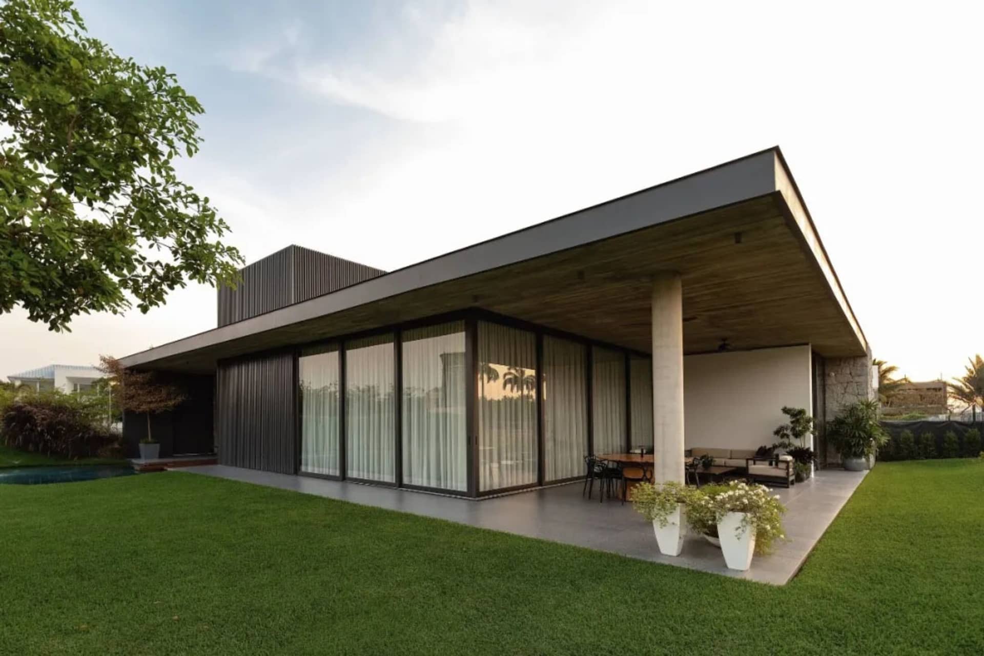 Modern residential house featuring glass windows, concrete pillars, metal roofing, and potted plants.