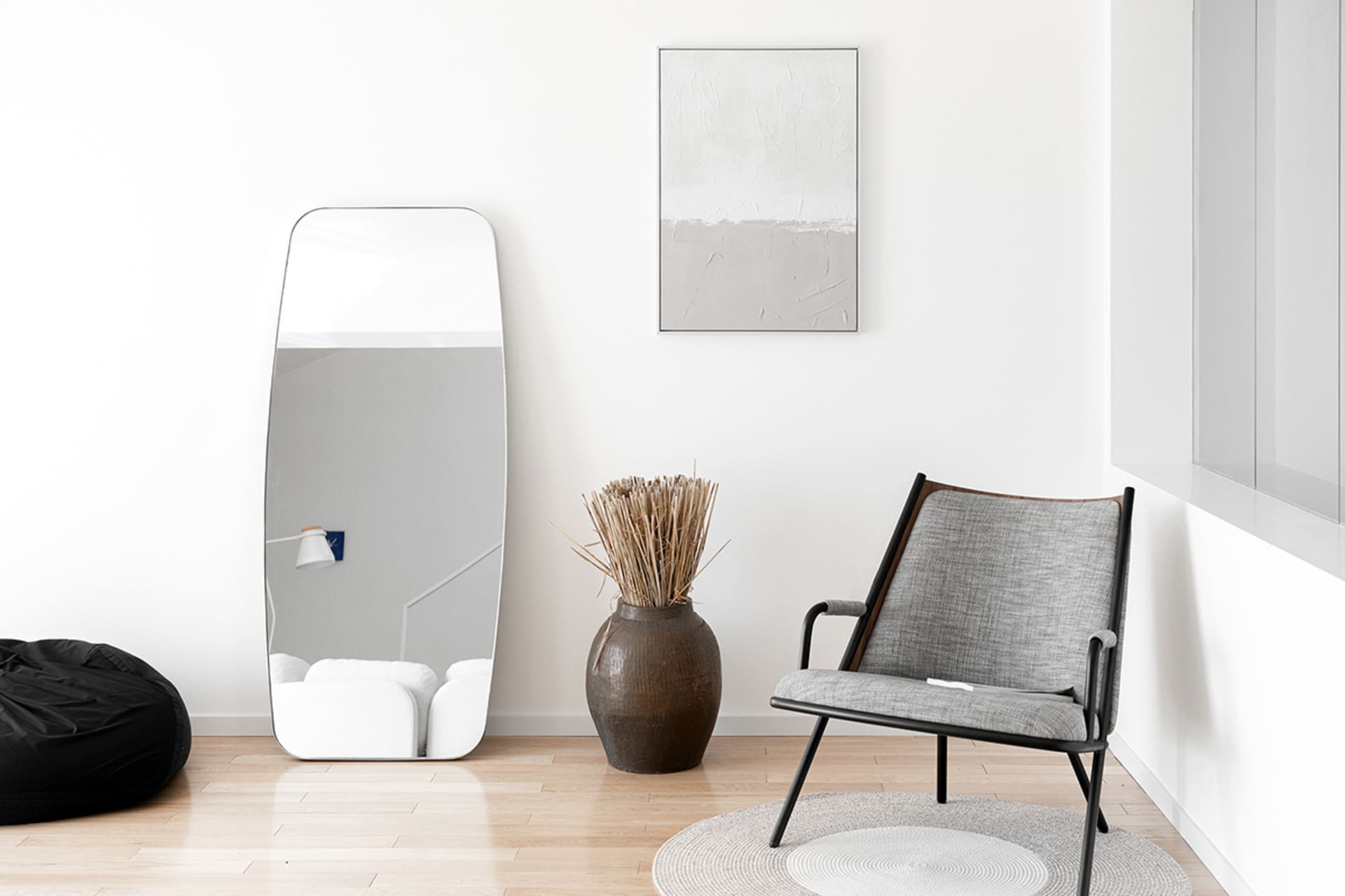 Modern minimalist living room with a gray upholstered chair, a large mirror, a vase with dried plants, and a black ottoman on