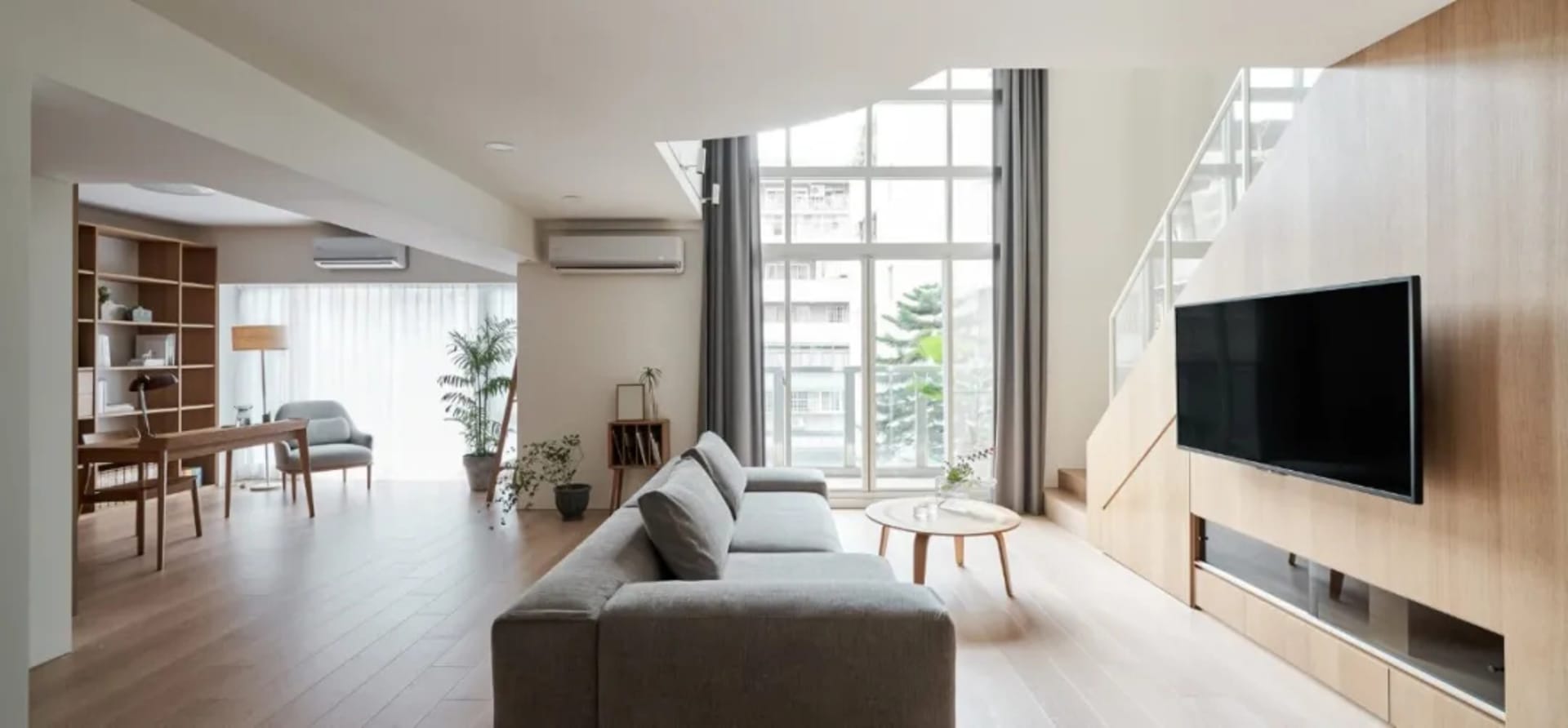 A modern minimalist living room with a grey couch, wooden floor, and large windows.