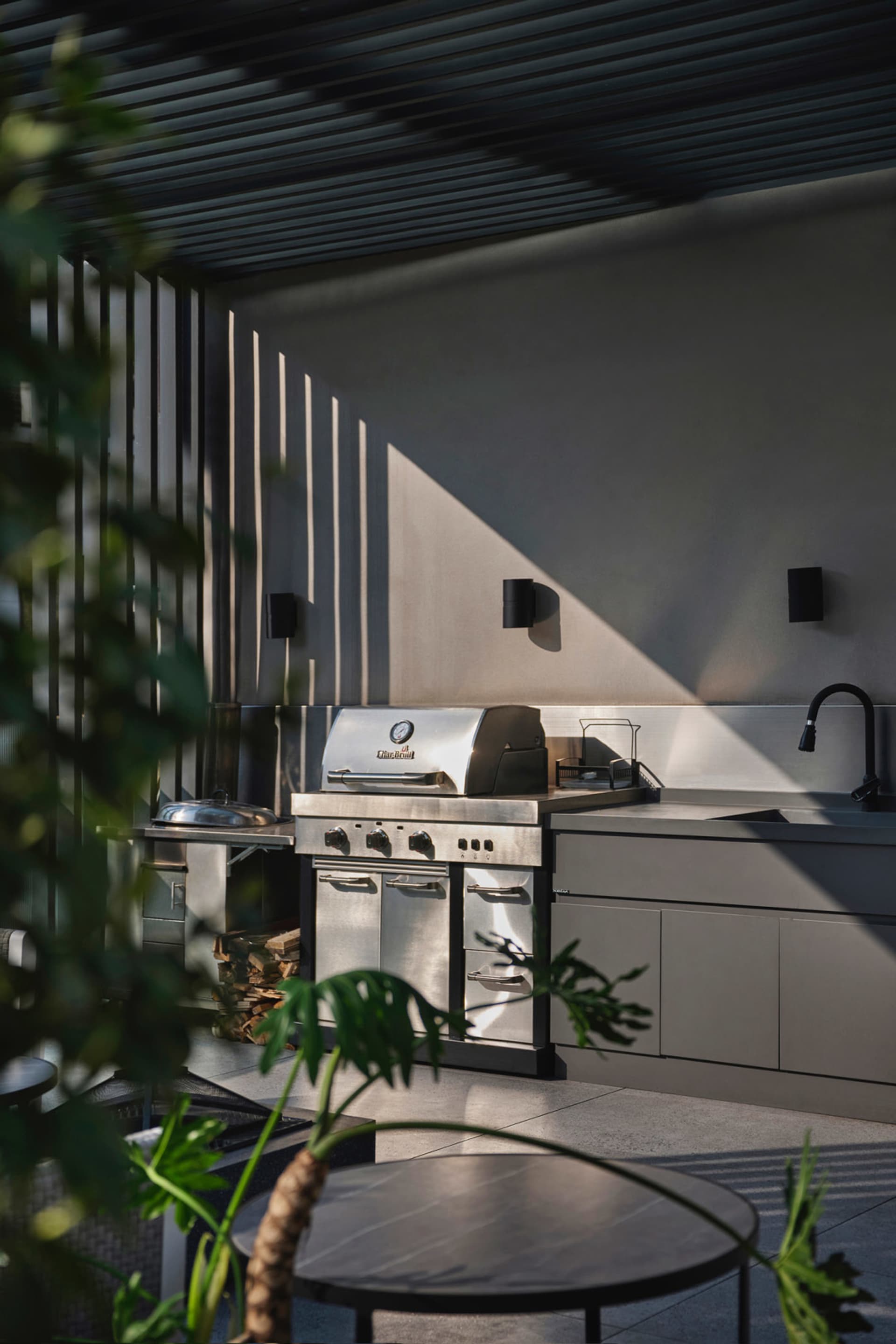 Modern, sleek outdoor kitchen with stainless steel appliances, concrete countertops, and a firewood storage area.