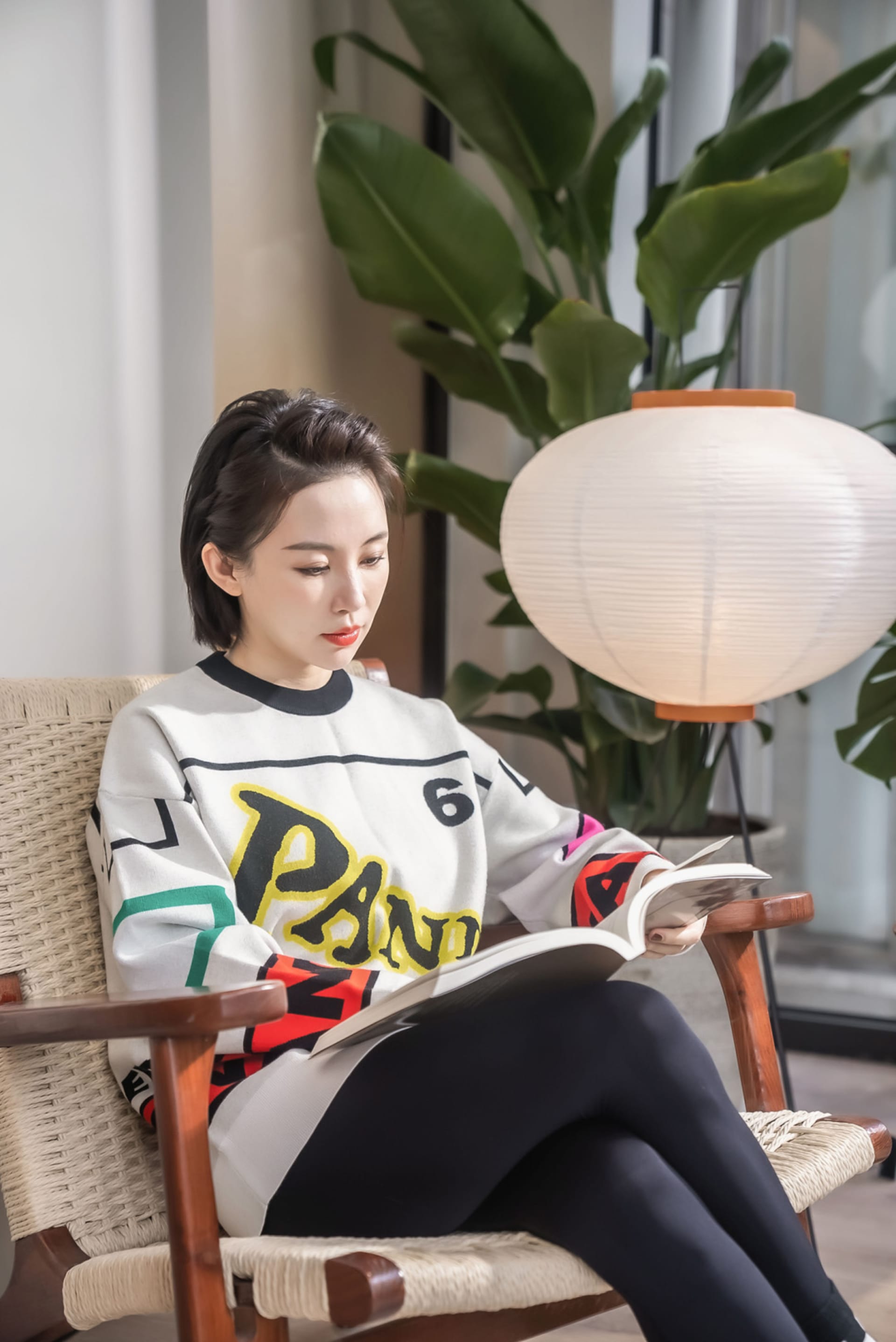 一位身穿彩色毛衣與黑色緊身褲的女子,坐在編織柳條扶手椅上閱讀書籍,旁邊擺放盆栽與落地燈。