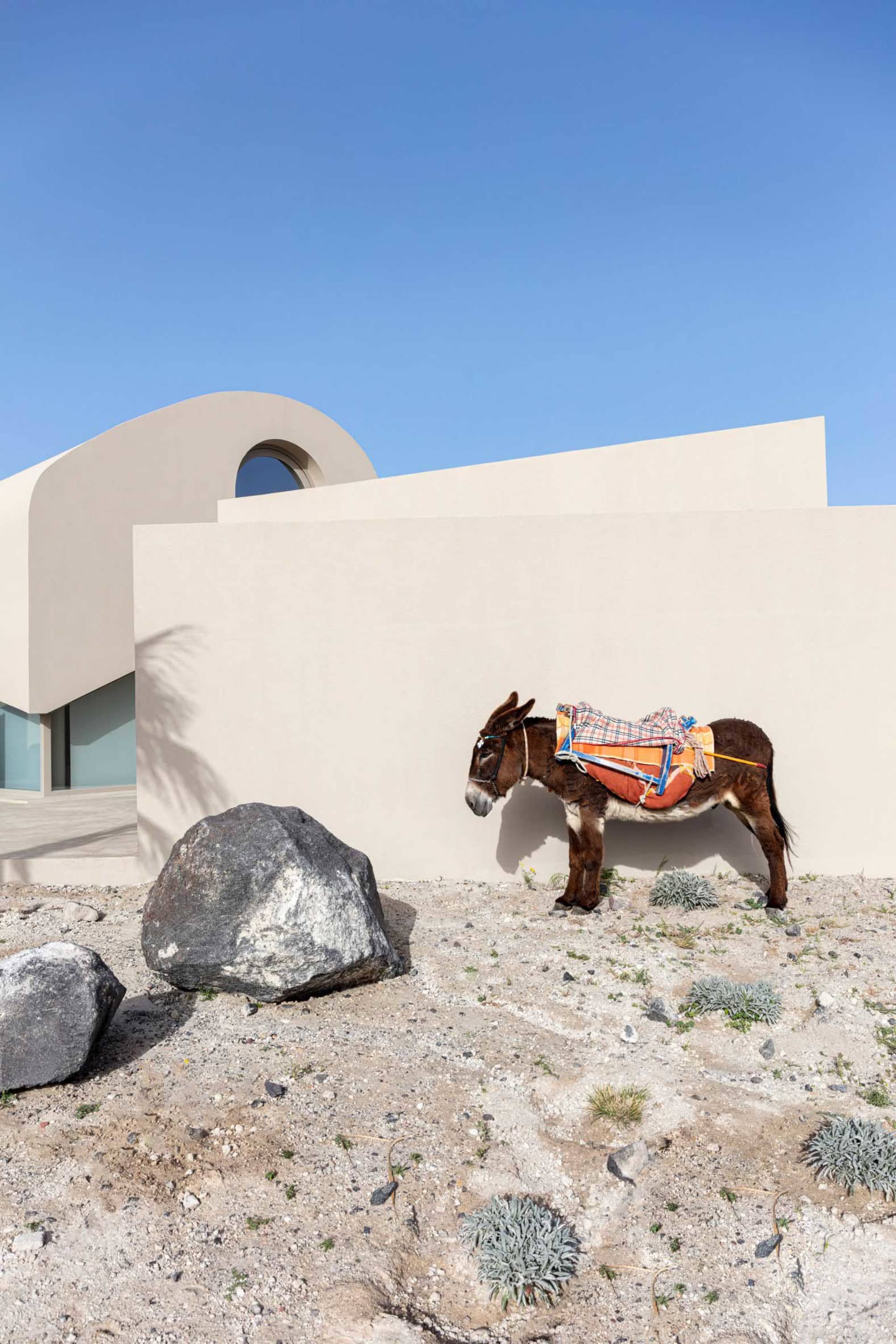 A minimalist house in a desert setting with a rock in the foreground, featuring sandstone walls and a circular window.