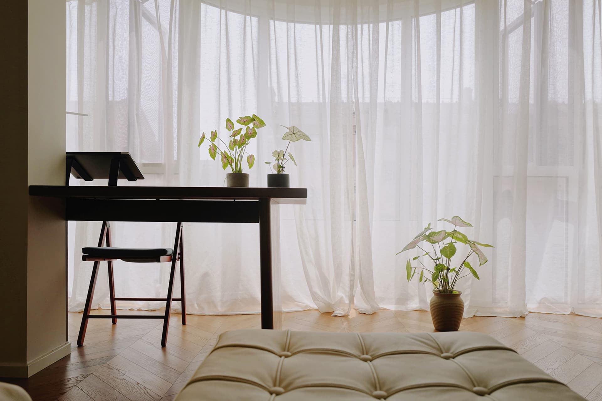 A cozy home office featuring a desk, chair, and potted plants under white curtains.