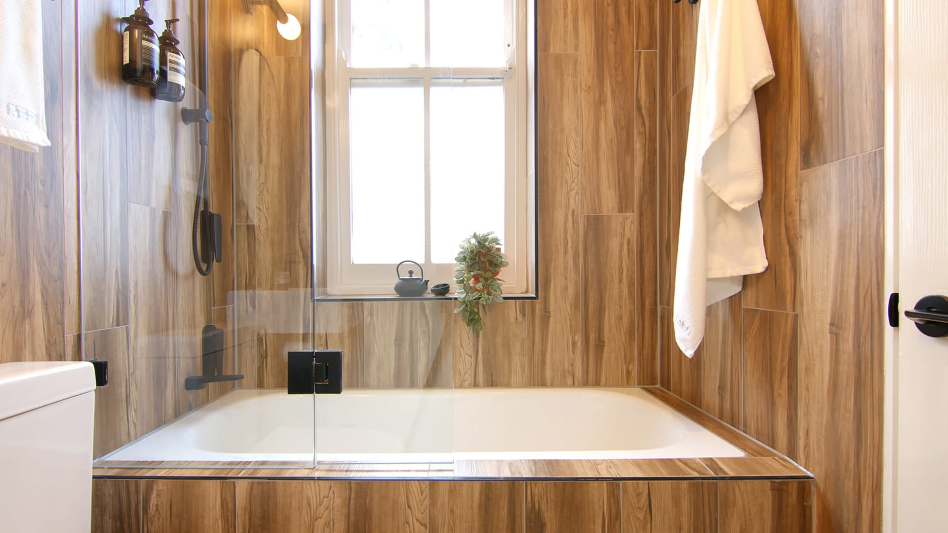 A modern bathroom with wood paneling, a glass-enclosed bathtub, and a window