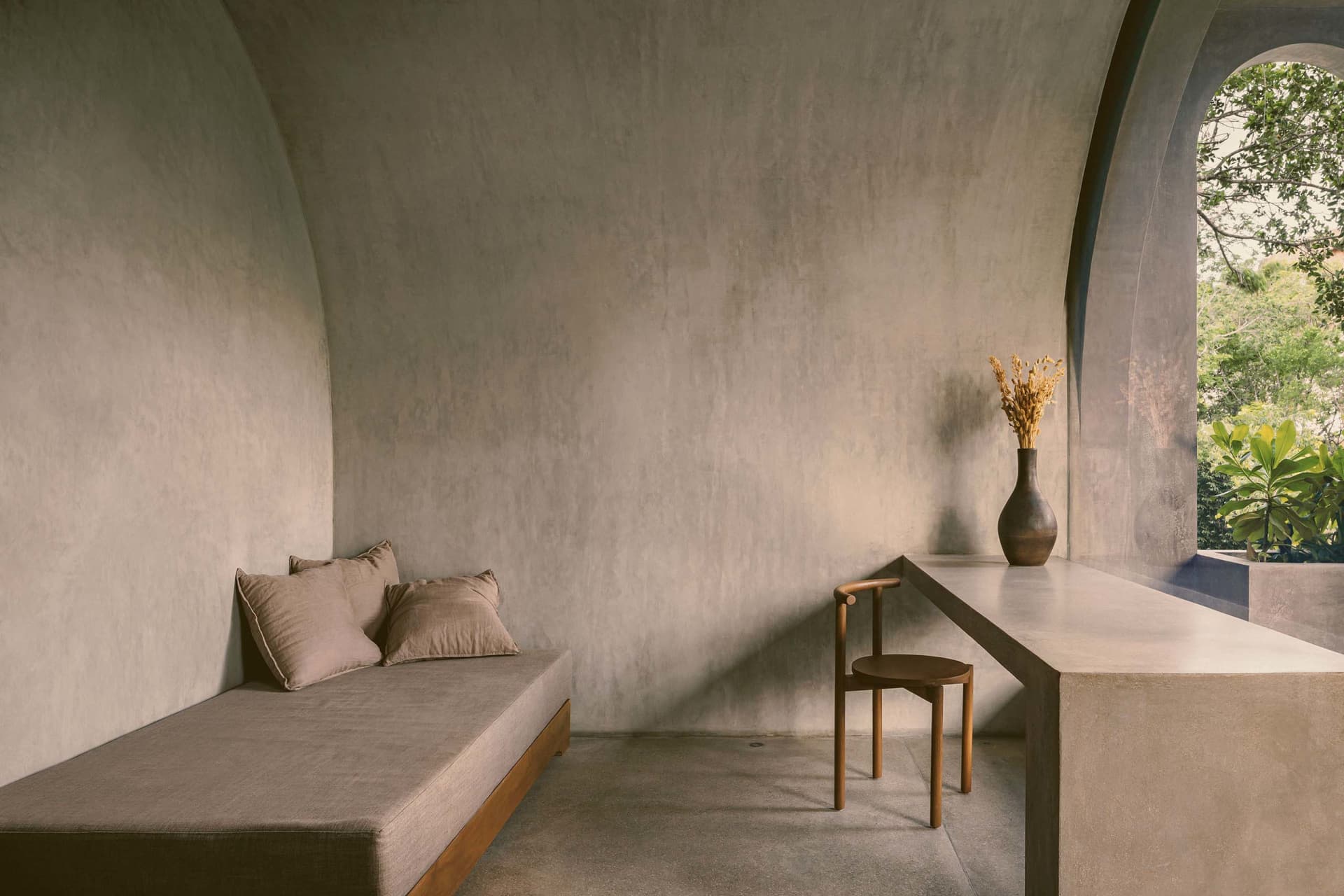 Modern minimalist living room with textured concrete walls and furniture, featuring a simple wooden chair with a vase of drie