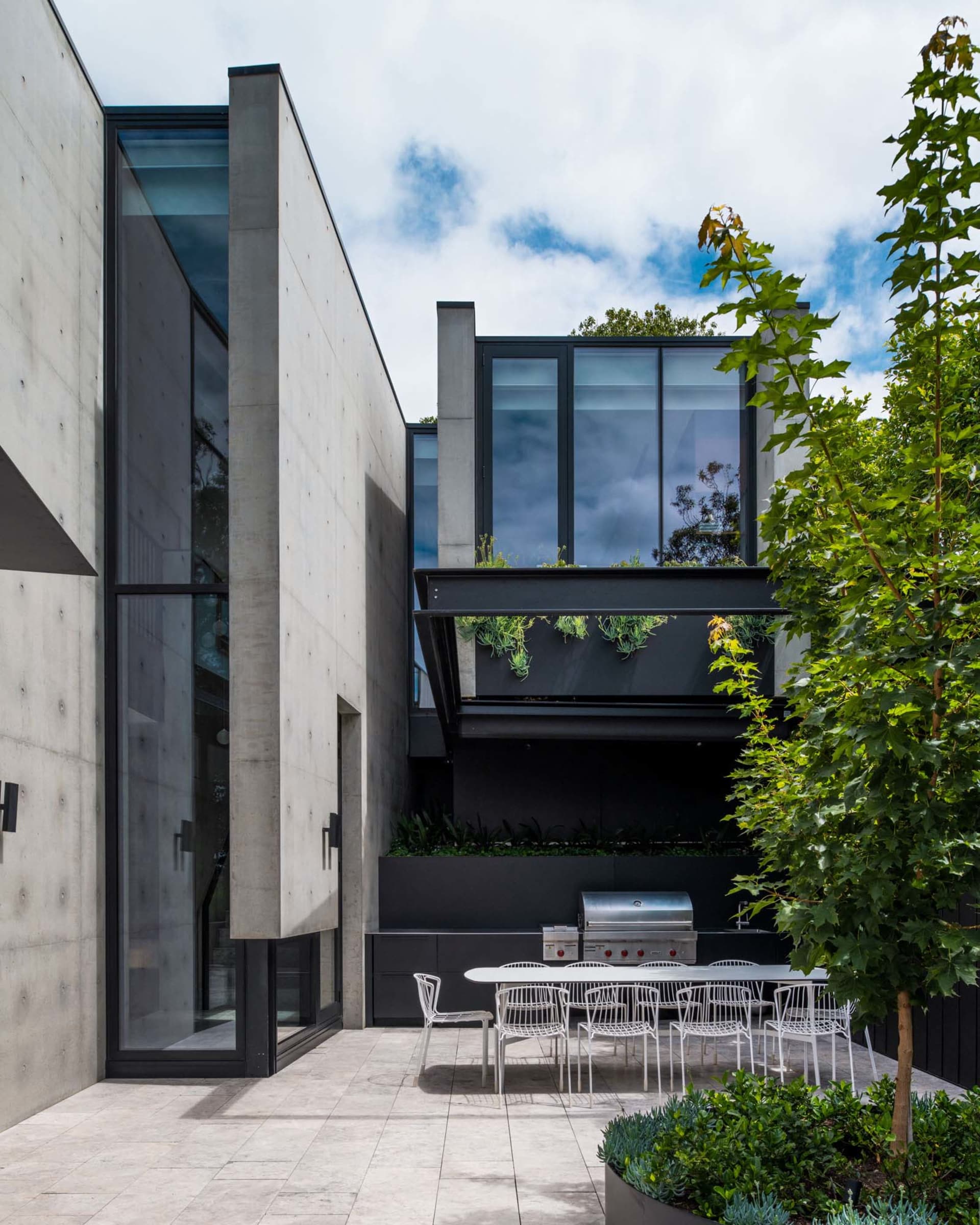 A modern residential patio featuring concrete flooring, glass walls, and a dining area with greenery and outdoor kitchen.