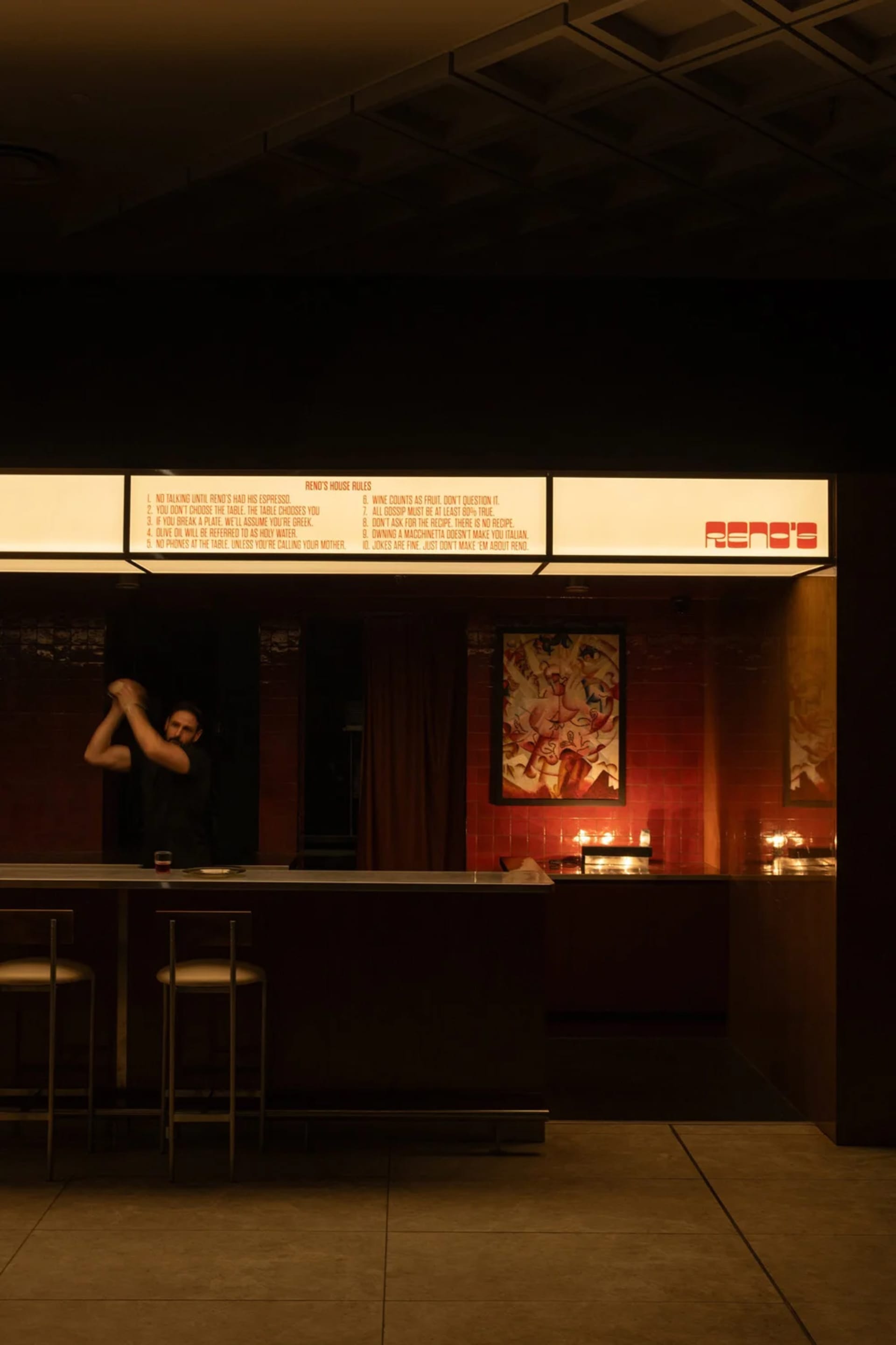 A casual bar with a brick wall, two chairs, and a counter with wine glasses, a cup, and a bowl, with a man standing behind the counter