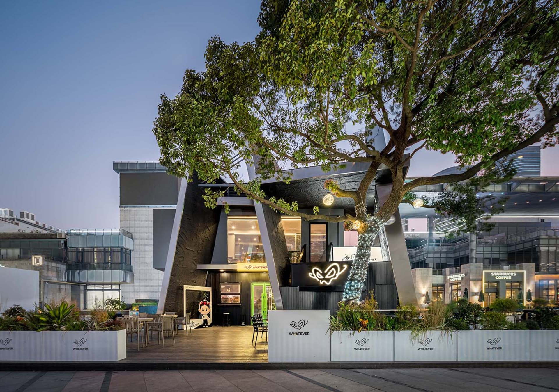 A modern building with a black facade and white accents, surrounded by a natural environment with a large tree and outdoor seating areas