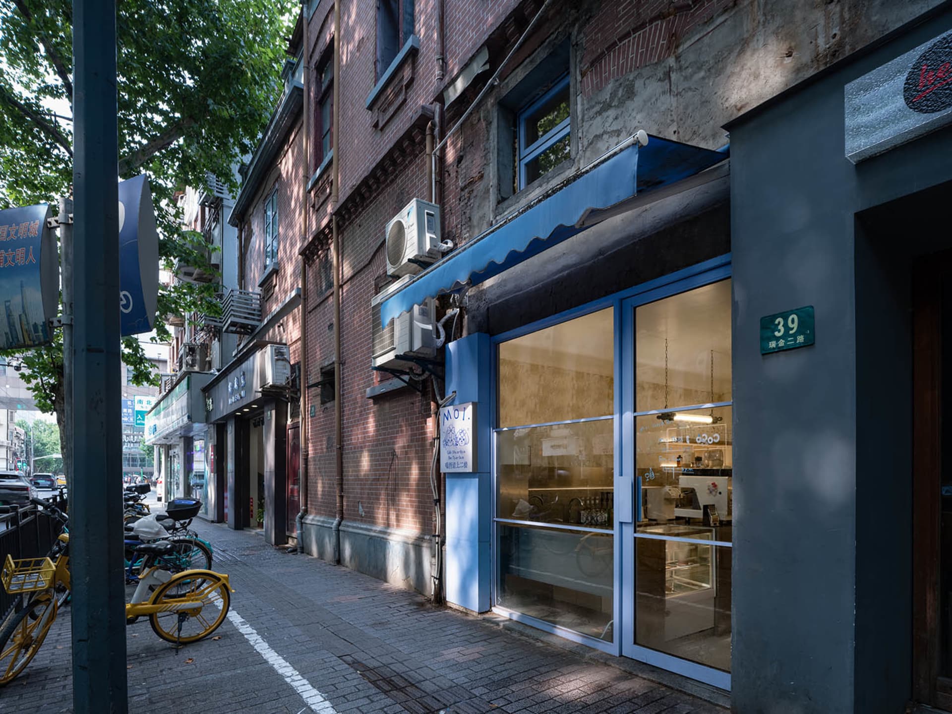 A street scene with a small cafe storefront, blue awning, bicycles, and a clock on the wall, with cups and possibly recent customer activity
