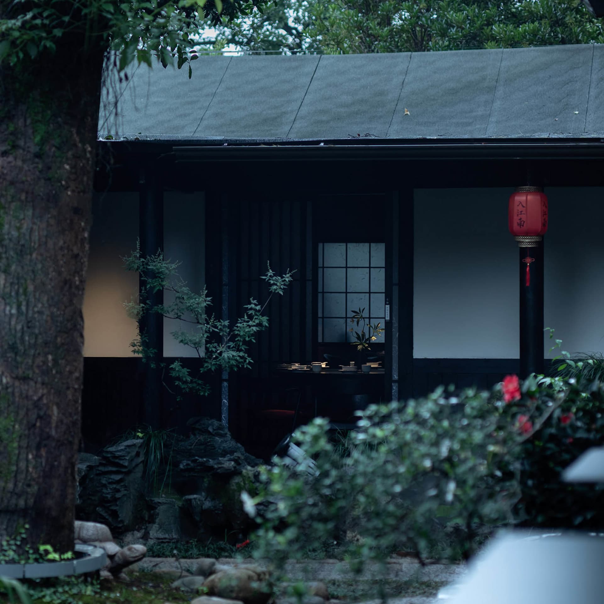 Asian-style house with a black exterior, surrounded by greenery, featuring a garden area with a rock wall and a potted plant near the entrance, with a peaceful and serene atmosphere