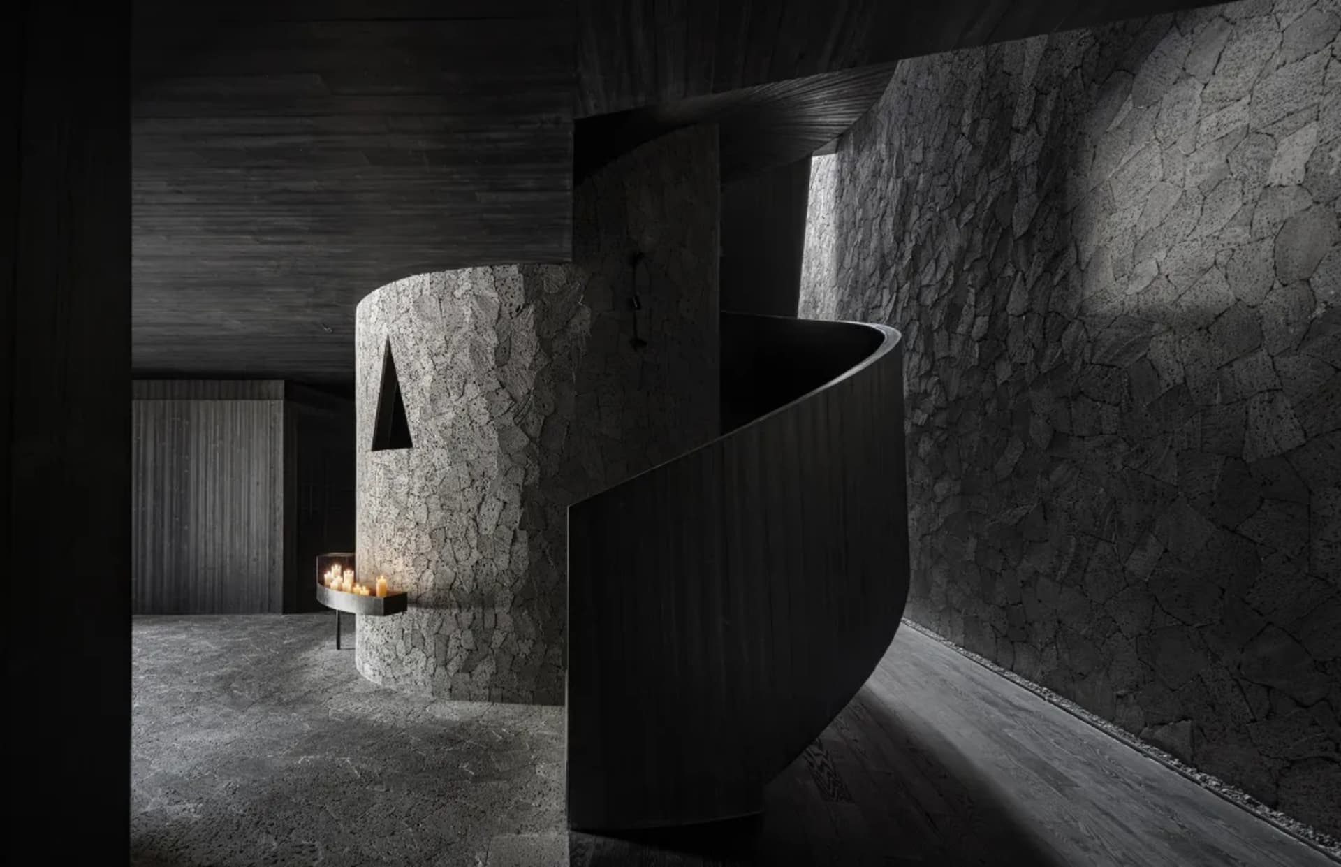 A modern living space with a stone spiral staircase, cave-like walls, a fireplace, and minimalist decor