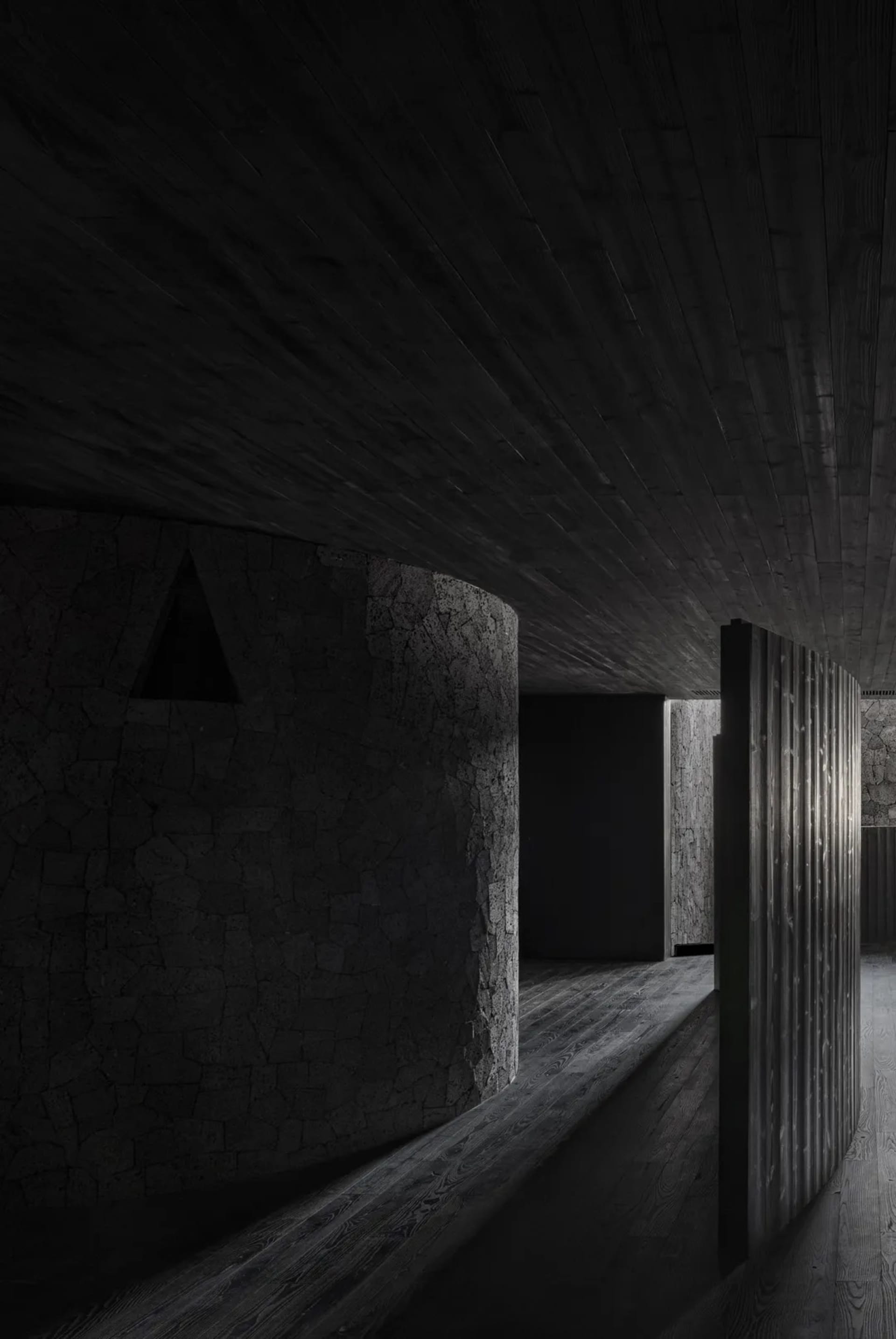 A large empty hallway with stone walls and a wooden floor, featuring two doorways and a darkened atmosphere