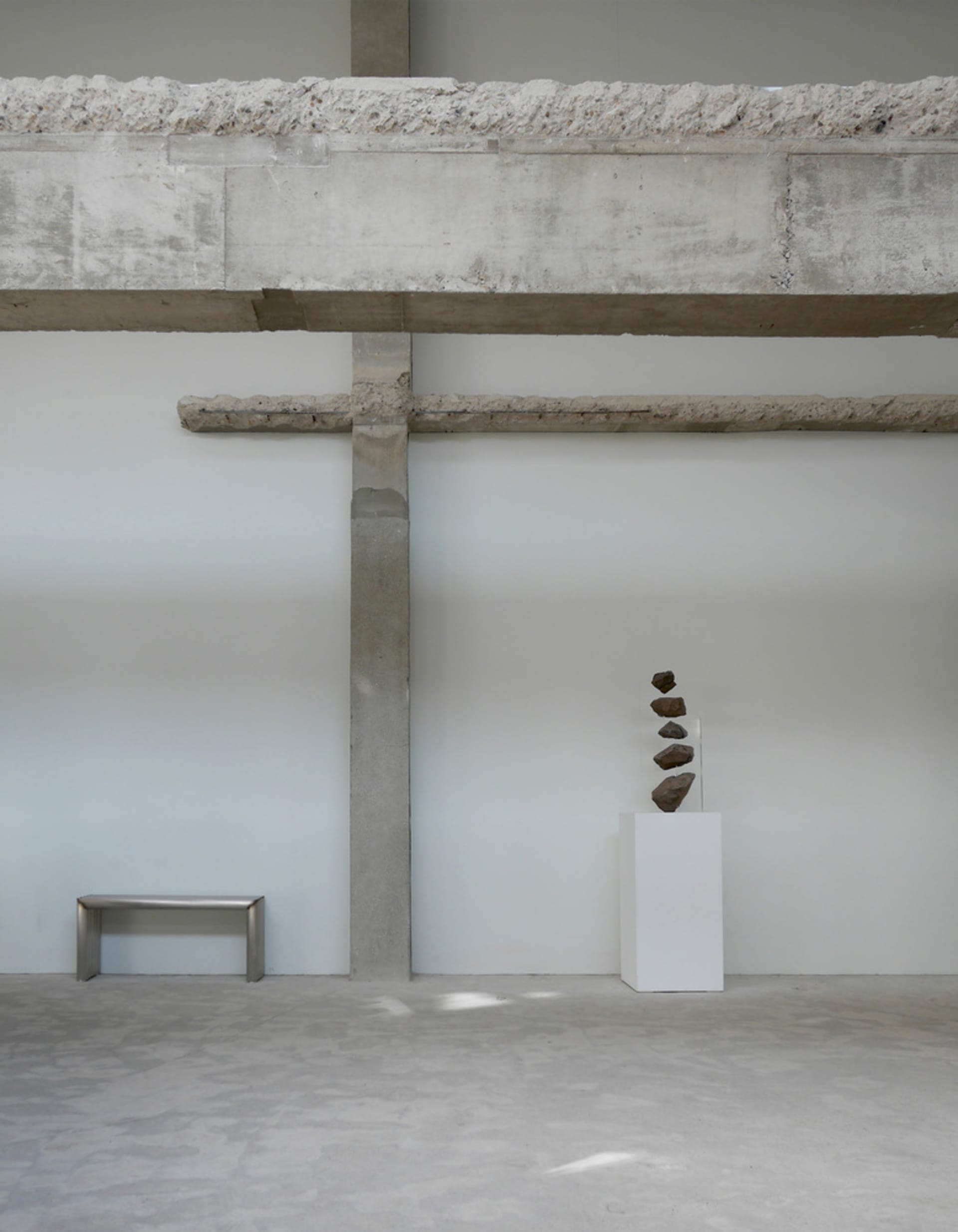 Empty industrial space with concrete walls and floors, featuring three sculptures and a bench, with a minimalistic and stark atmosphere