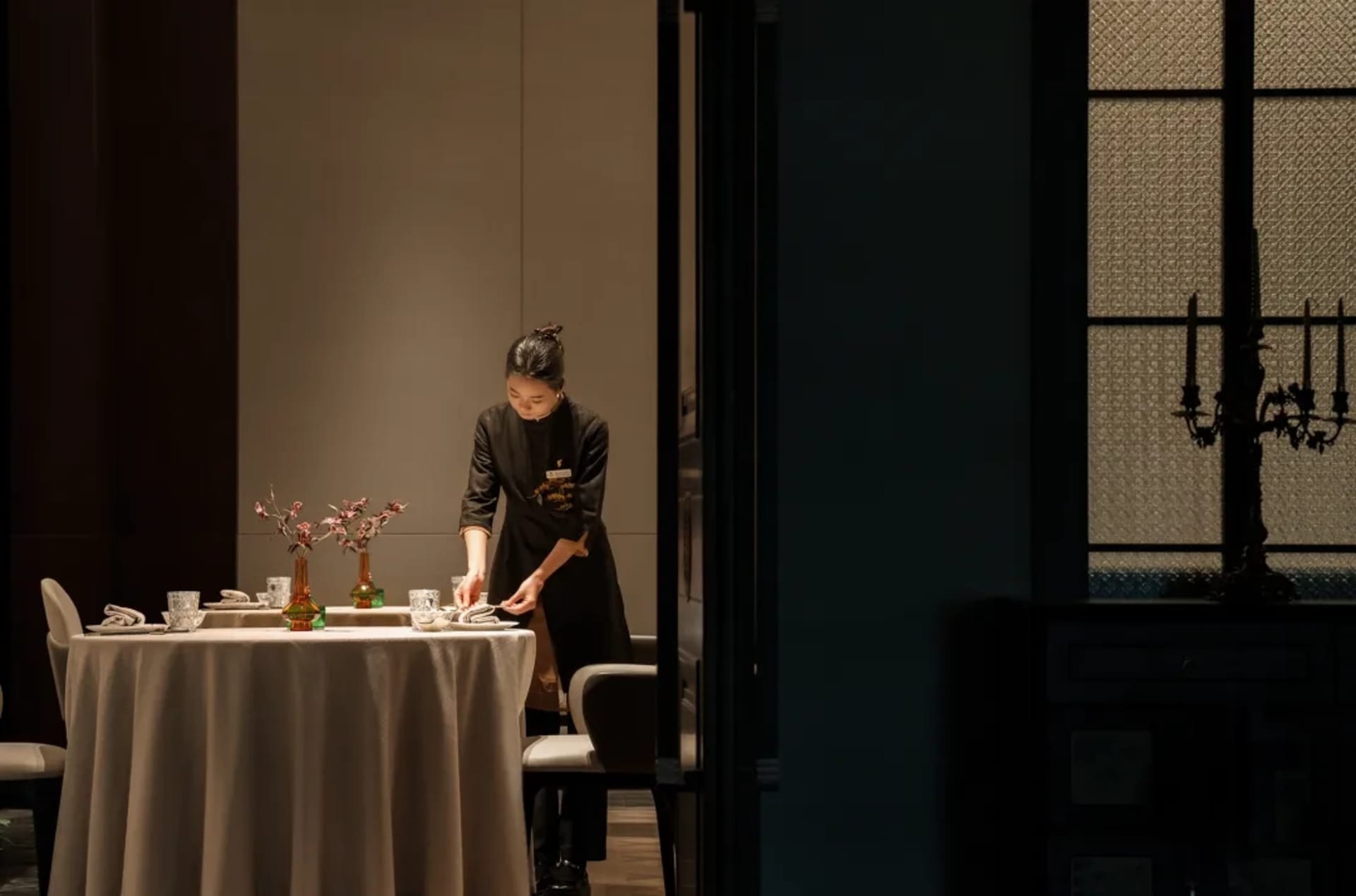 A woman in an apron prepares food on a table in a professional kitchen with bowls, a spoon, and a knife