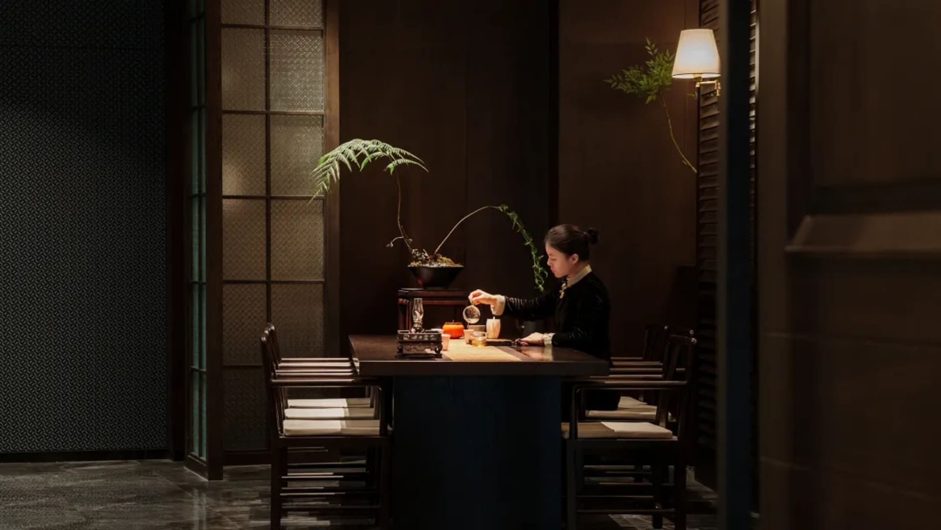 一位女性坐在餐桌旁，置身於燈光昏暗的房間，周圍環繞著椅子，營造出溫馨親密的氛圍