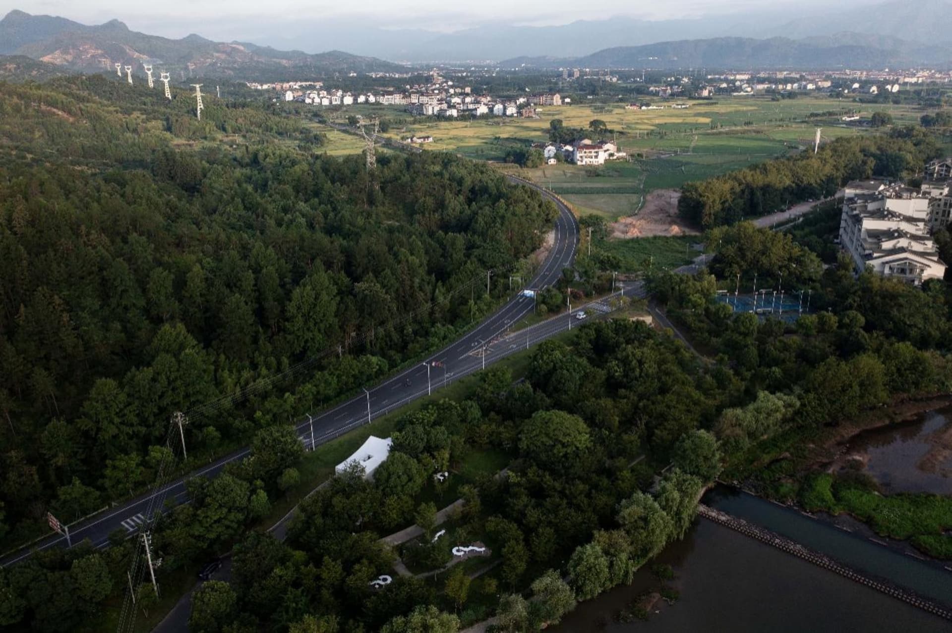 Aerial view of a residential area with houses, trees, and roads, surrounded by greenery and featuring parked cars
