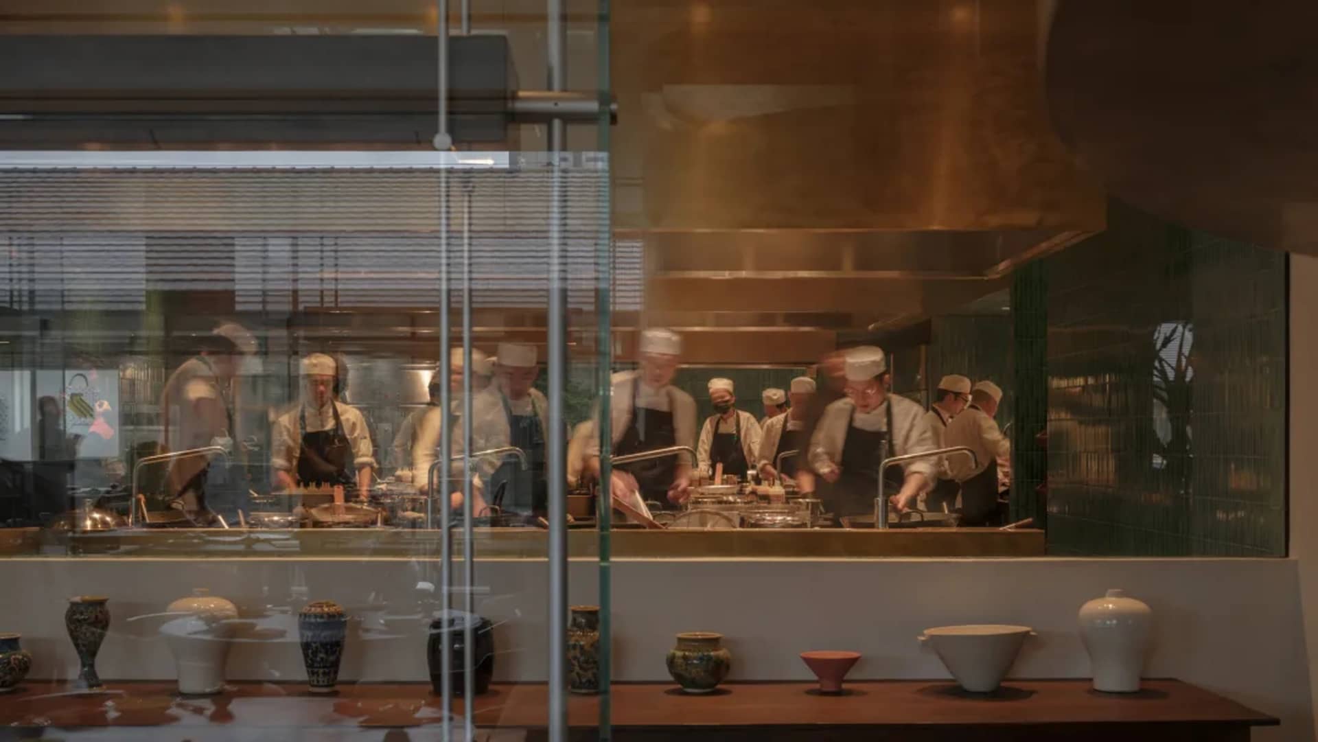 A bustling commercial kitchen with multiple chefs preparing meals, featuring various bowls and cups scattered throughout the area