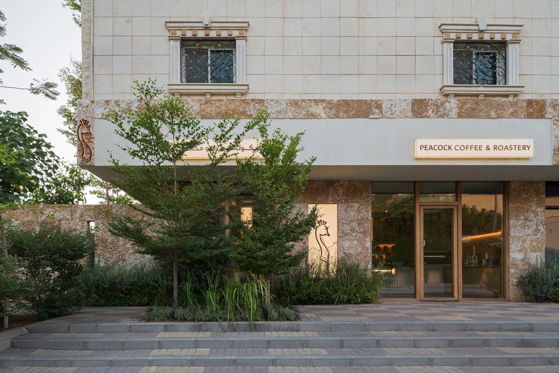 A stone building facade with a large window, surrounded by two trees in a serene environment