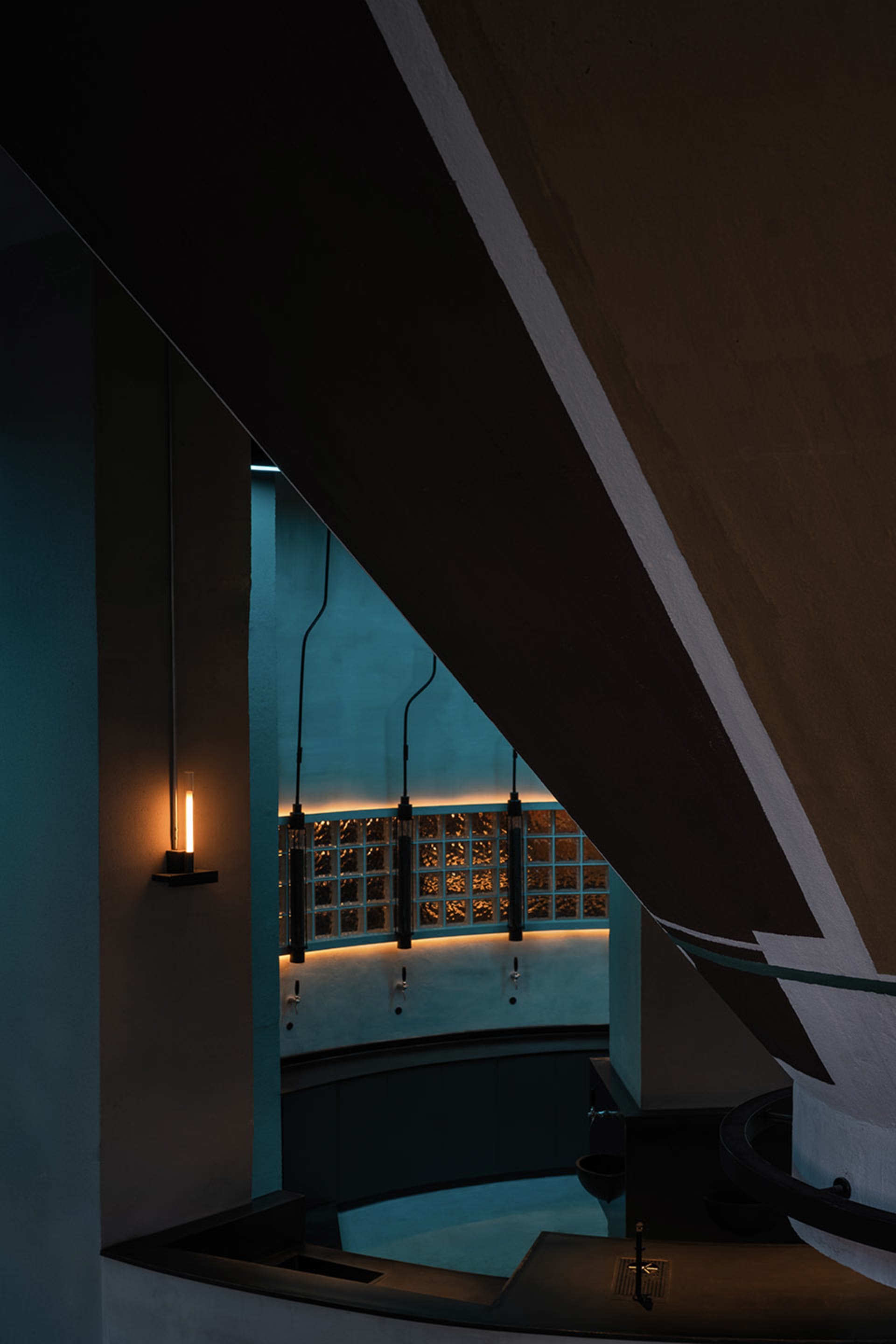 A wooden staircase with a traditional appearance, featuring a wooden railing on both sides, illuminated by two lights, one near the bottom left and another towards the center right
