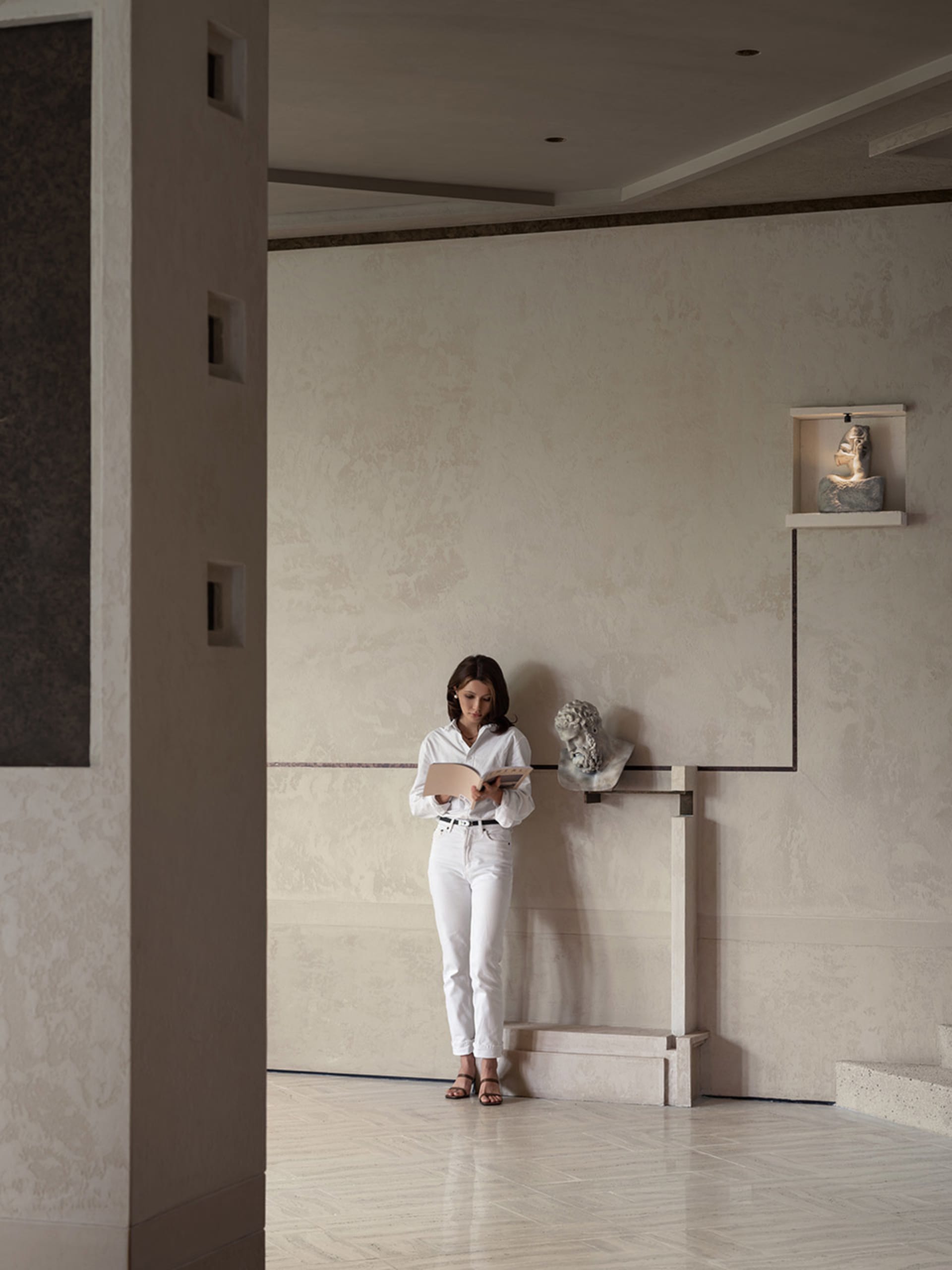 A woman reads a book in a calm, empty room with a white color scheme