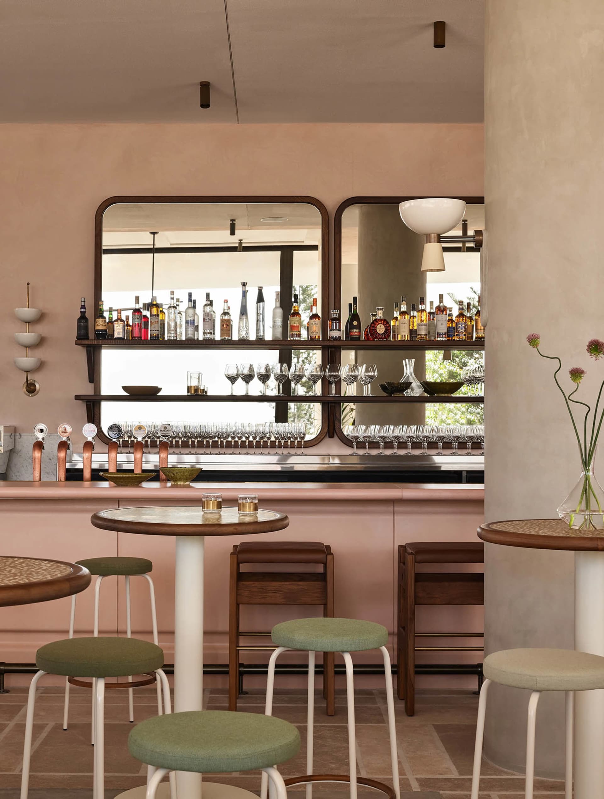 A pink-themed bar area with a long counter, wine glasses, and chairs, surrounded by bottles and conveying a cozy atmosphere
