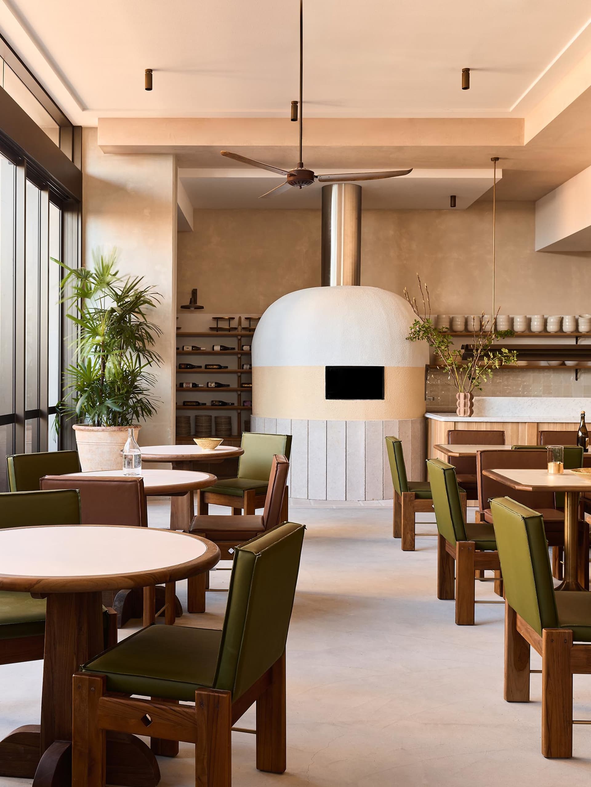 Cozy modern restaurant dining area with a stone oven, wooden tables, and vases, surrounded by chairs and warm lighting